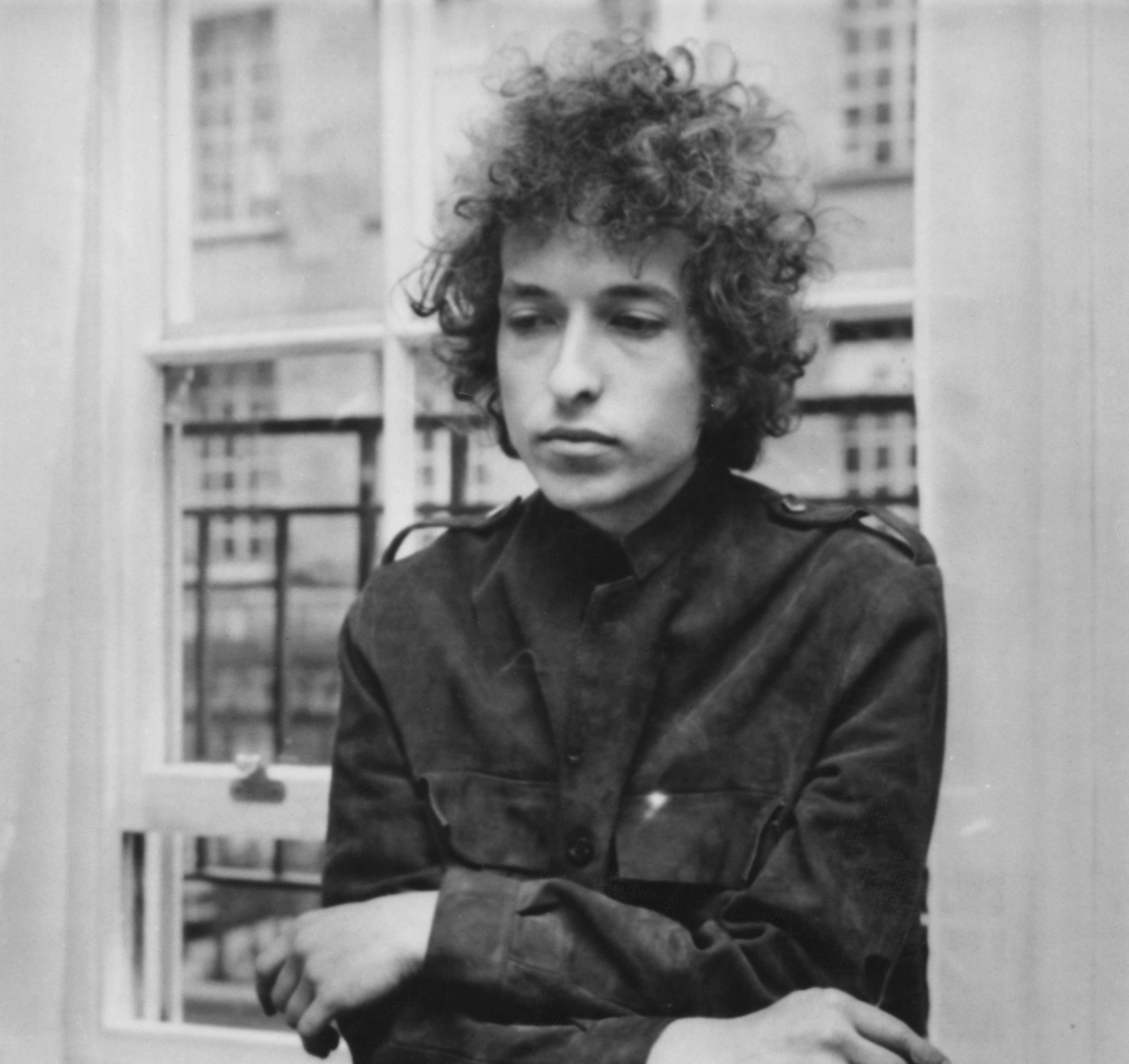 A black and white photo of Bob Dylan standing in front of a window. 