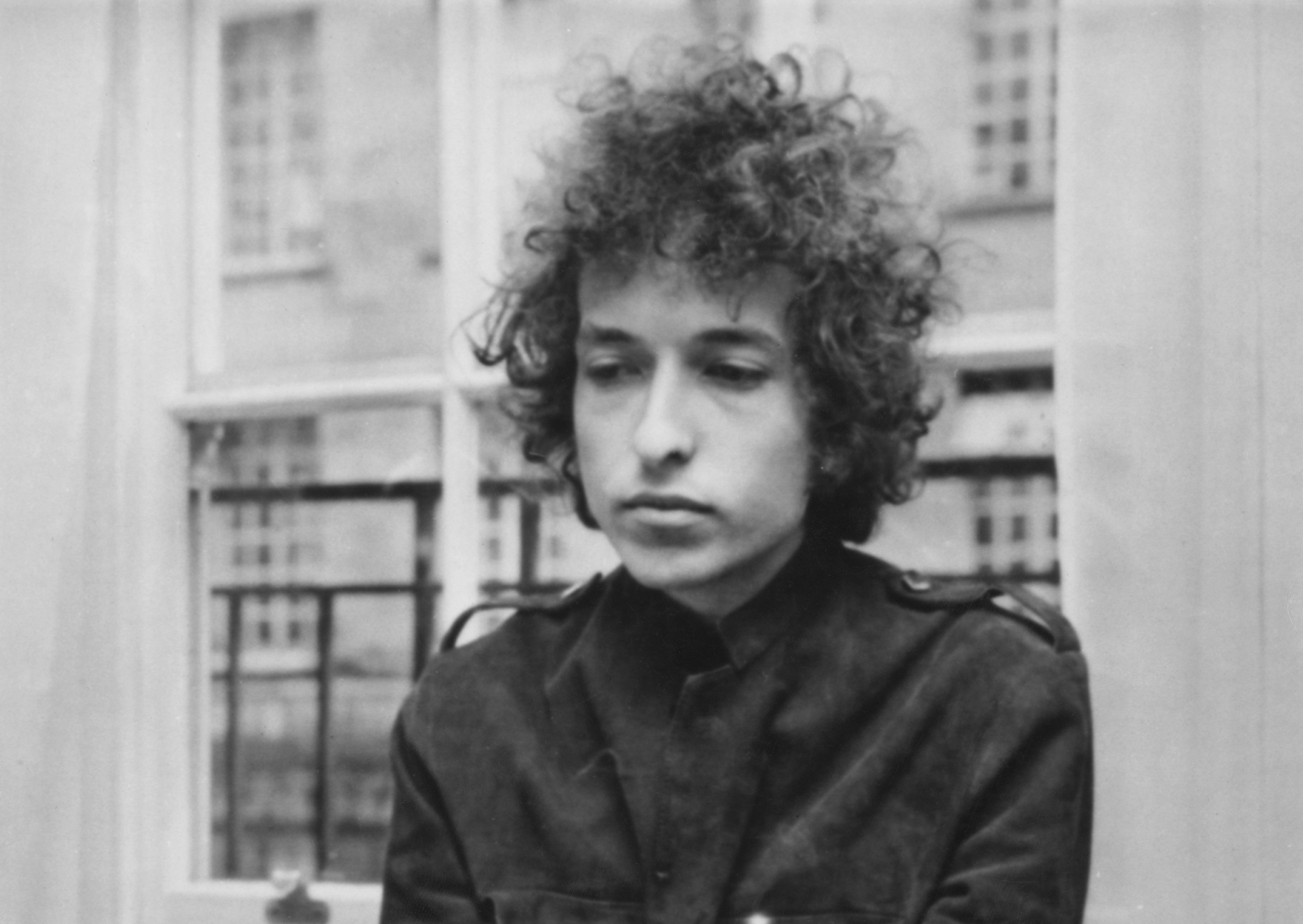 A black and white picture of Bob Dylan standing in front of a window.