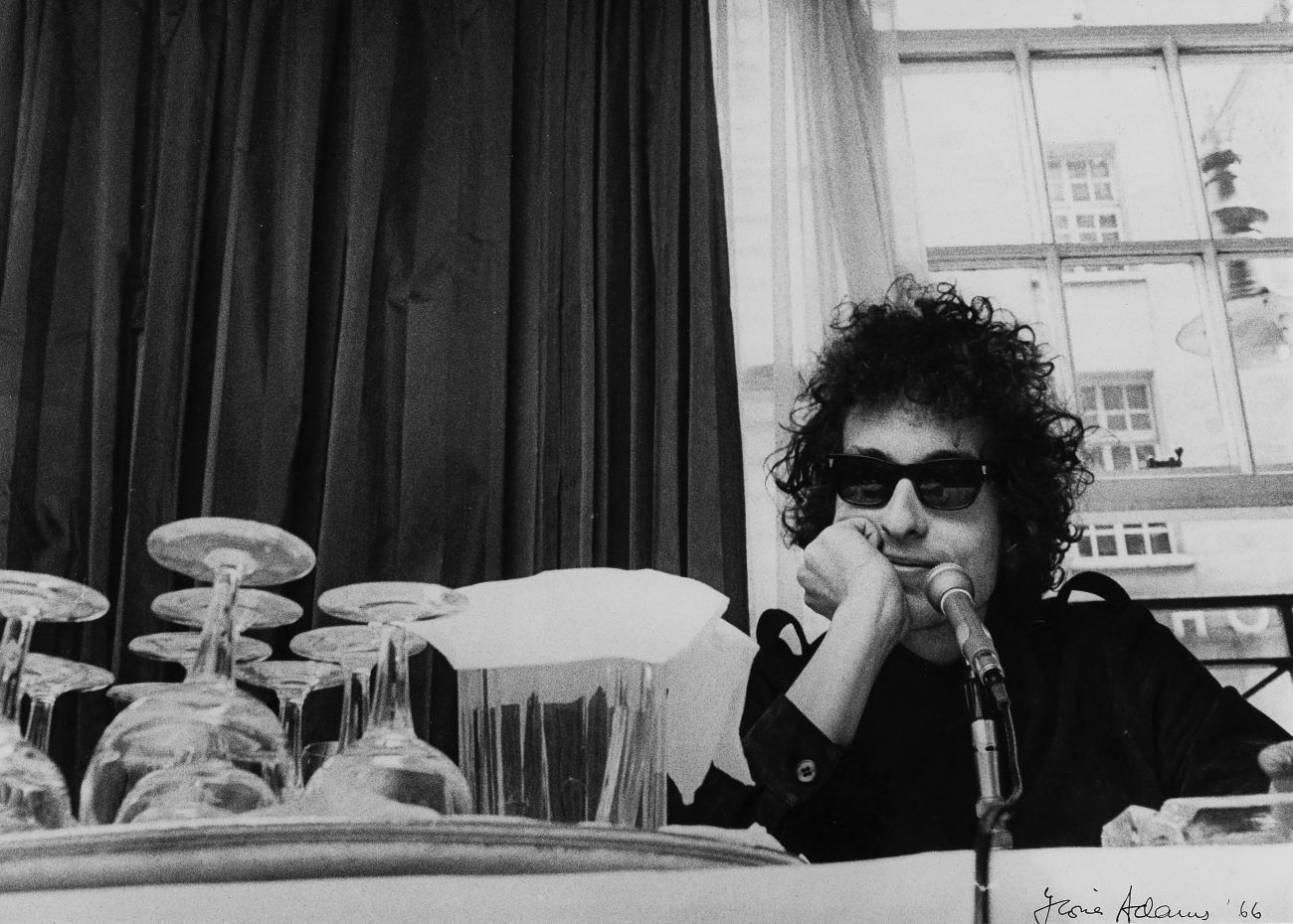 Bob Dylan wears sunglasses and sits at a table in front of a microphone. 