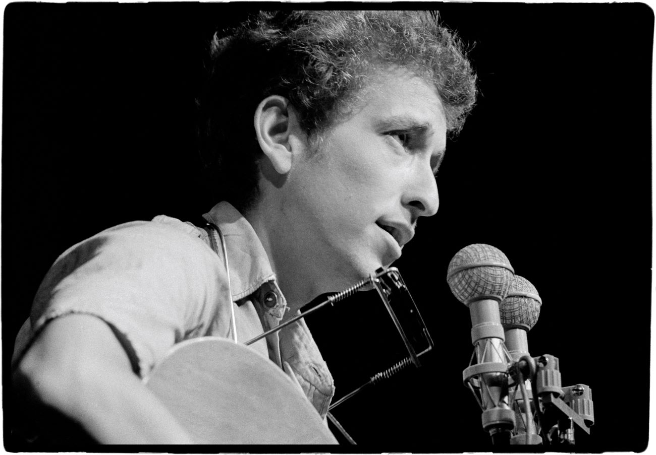 A black and white picture of Bob Dylan with a guitar and a harmonica, in front of a microphone.