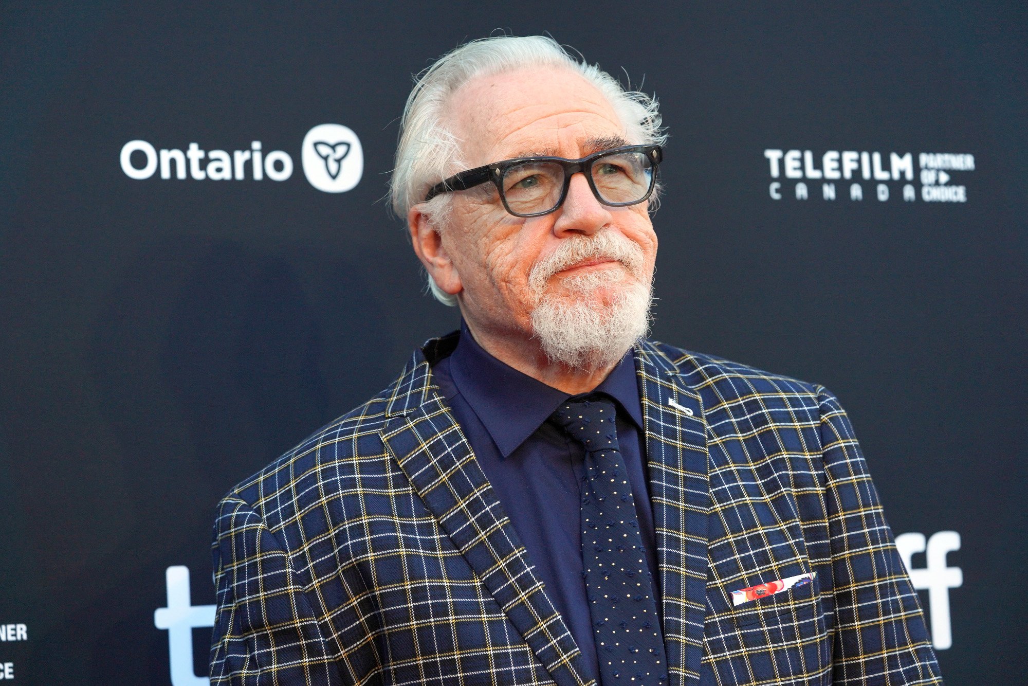 'Succession' cast member Brian Cox on the red carpet at the Toronto International Film Festival. He's wearing a dark blue shirt, blue and yellow checkered jacket, and glasses.