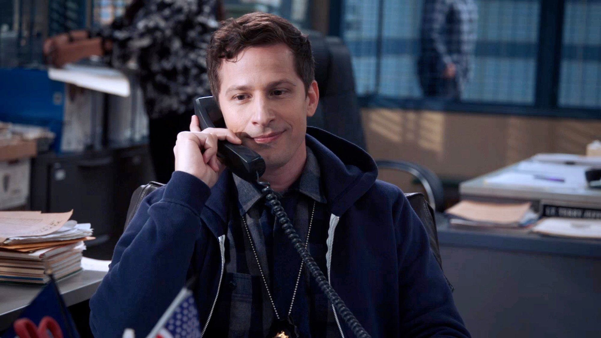 Andy Samberg holds a phone at a desk in a production still from 'Brooklyn Nine-Nine,' which is leaving Hulu in September 2022.