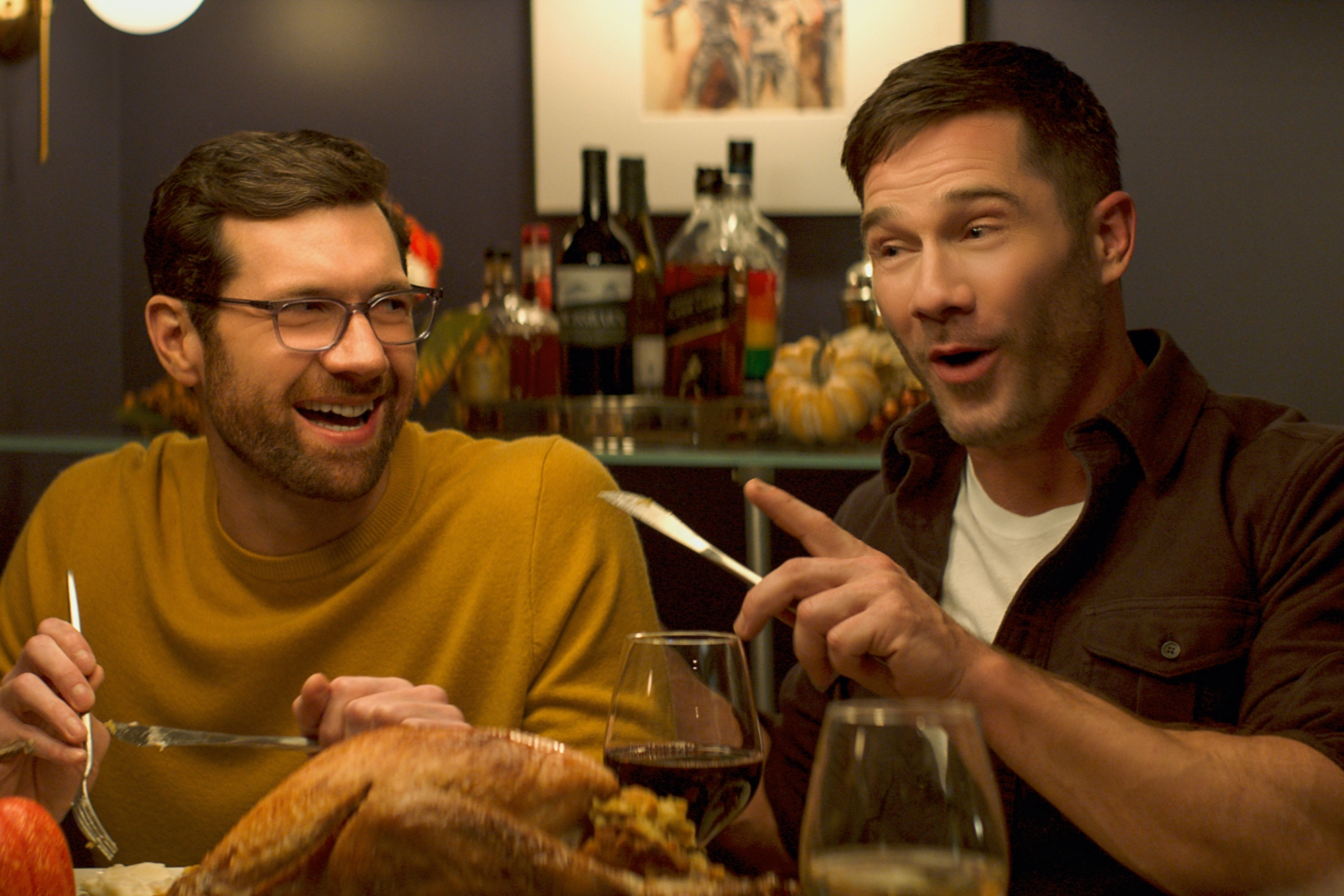 'Bros' Billy Eichner as Bobby Leiber and Luke Macfarlane as Aaron Shepard. Eichner is laughing wearing a yellow shirt holding a fork. Macfarlane is wearing a white shirt under an overshirt holding a butterknife and pointing across the table.
