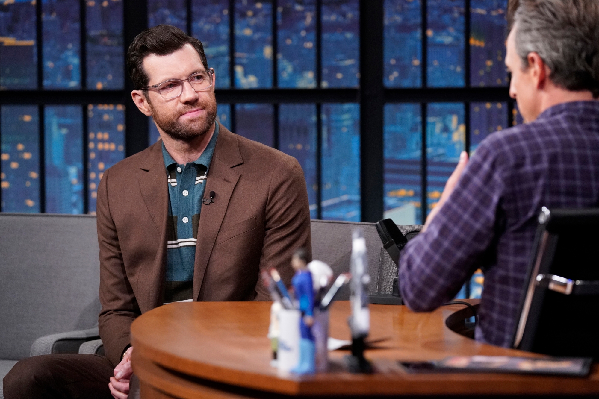 'Bros' actor Billy Eichner telling host Seth Meyers about Tinder bans. Eichner is wearing a brown suit jacket and glasses, looking at Meyers from the other side of the desk. Meyers is shown from the back wearing a plaid shirt.