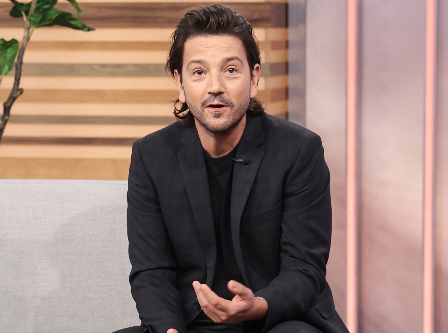 Cassian Andor actor Diego Luna. He's sitting, wearing a black suit, and speaking.