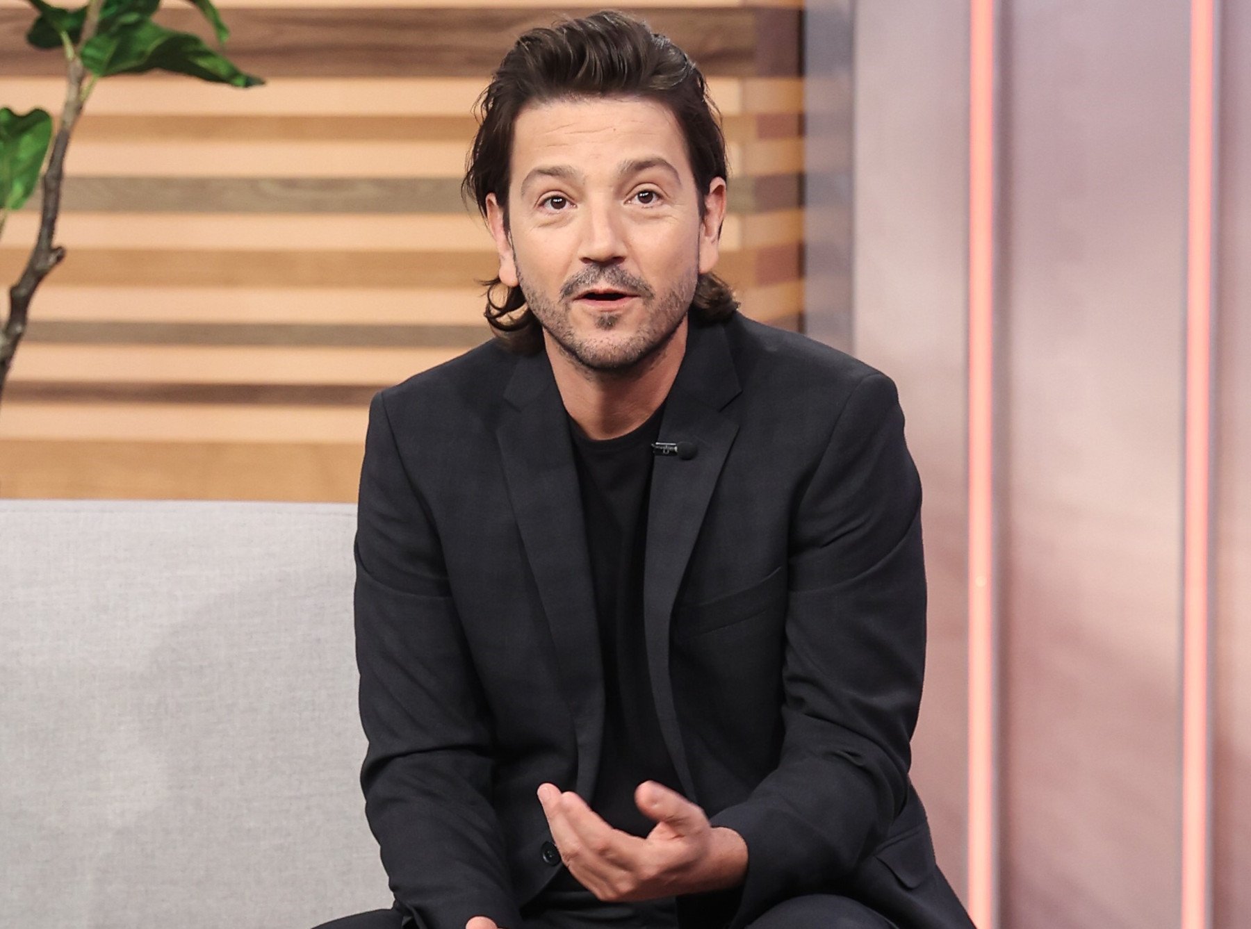 Cassian Andor actor Diego Luna. He is sitting, wearing a black suit and speaking.