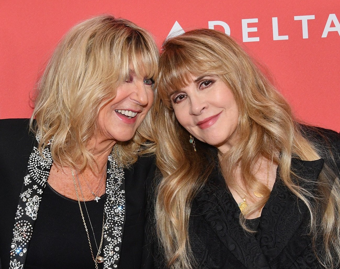 Stevie Nicks and Christine McVie stand with their heads together in front of a red background. 