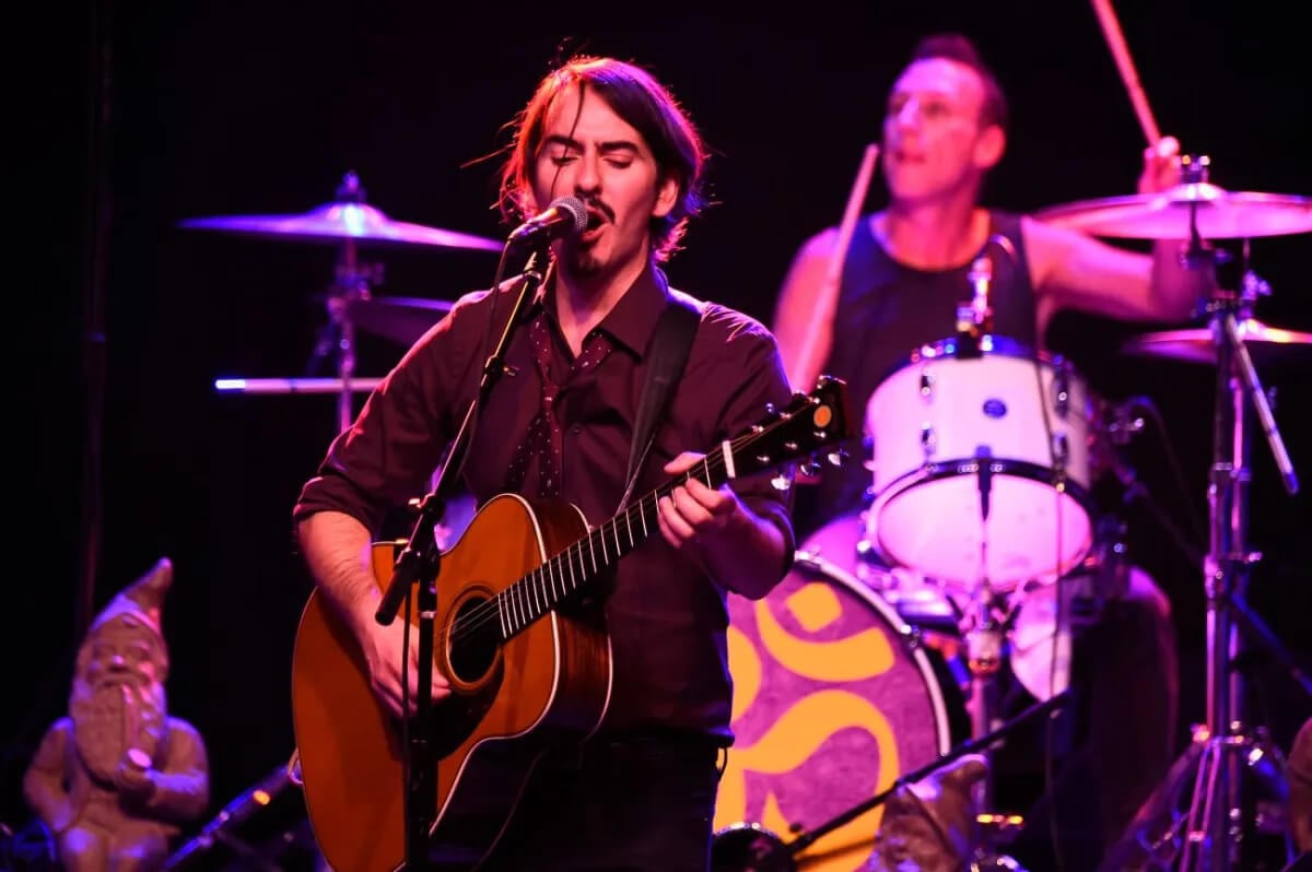 George Harrison's son Dhani plays the guitar and sings into a microphone.