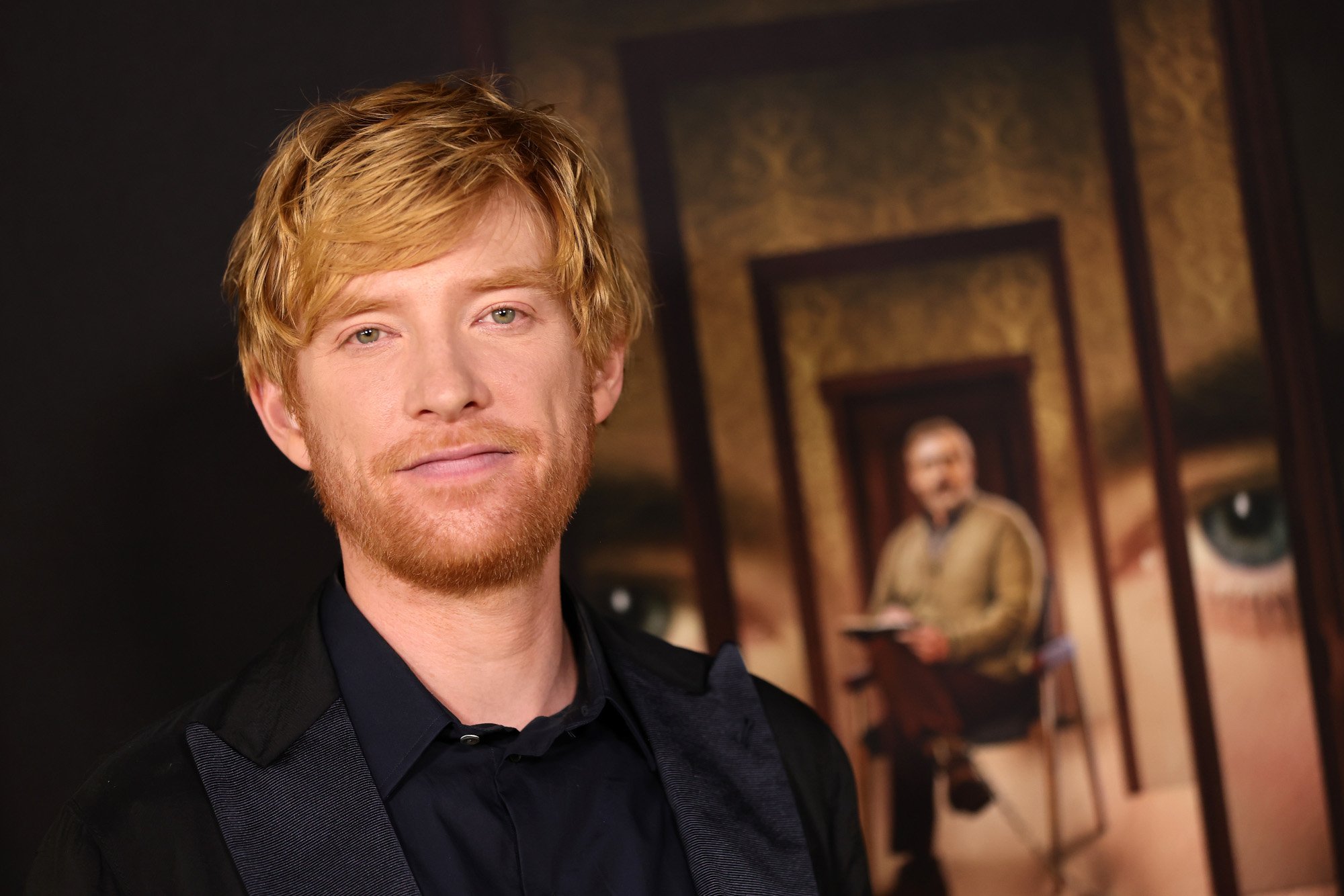 'The Patient' star Domhnall Gleeson. He's wearing a black shirt, black jacket, and standing in front of a poster for the Hulu series.