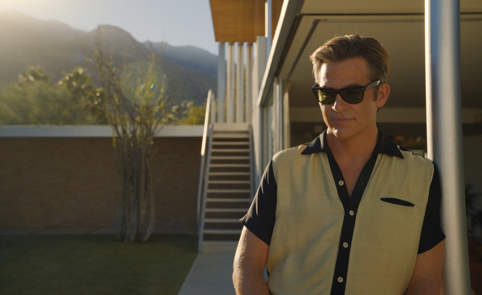 'Don't Worry Darling' Chris Pine as Frank. He's wearing sunglasses and a collared shirt and leaning against his modern home with the external staircase in the background.