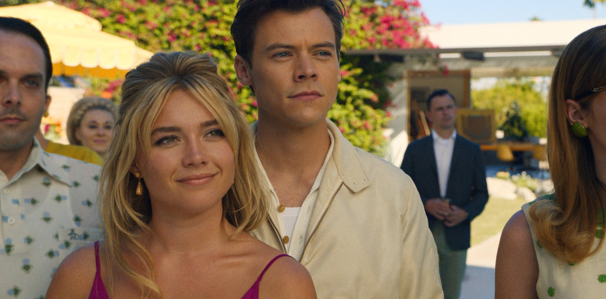 'Don't Worry Darling' Florence Pugh as Alice Chambers and Harry Styles as Jack Chambers. Pugh is smiling wearing gold earrings and a purple top. Styles is standing next to her slightly smiling wearing off-white colored clothes, surrounded by their friends.