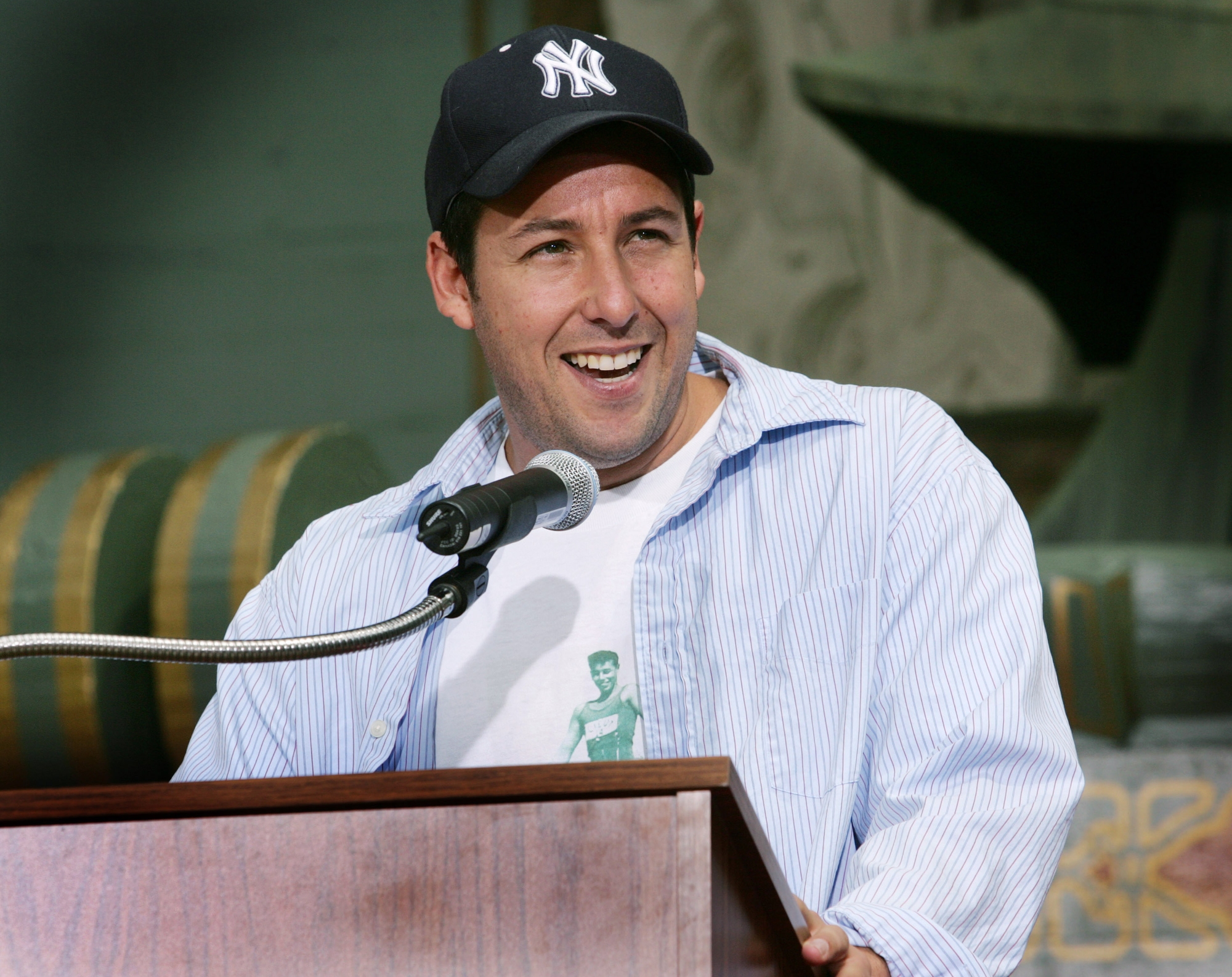 Ex-'Saturday Night Live' cast member Adam Sandler standing behind a podium with a microphone. He's smiling and wearing a baseball cap and an opened collared shirt with a white shirt underneath.
