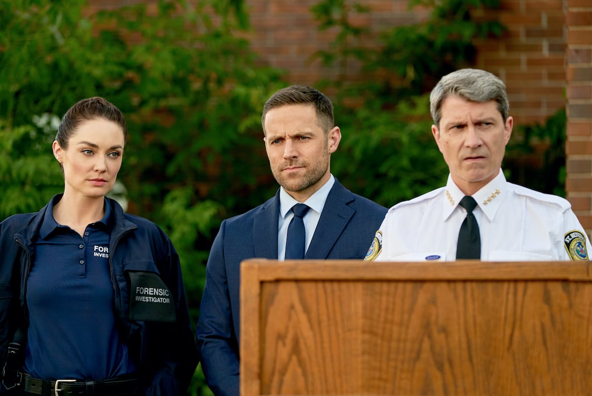 Mallory Jansen and Dylan Bruce standing next to a man behind a lectern in the Hallmark mystery movie 'Francesca Quinn, P.I.'