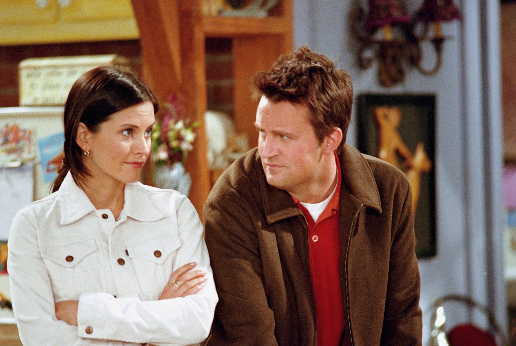 'Friends' Courteney Cox as Monica and Matthew Perry as Chandler. Monica is wearing a white jacket and crossing her arms. Chandler is wearing a brown jacket and a red shirt underneath looking at Monica.