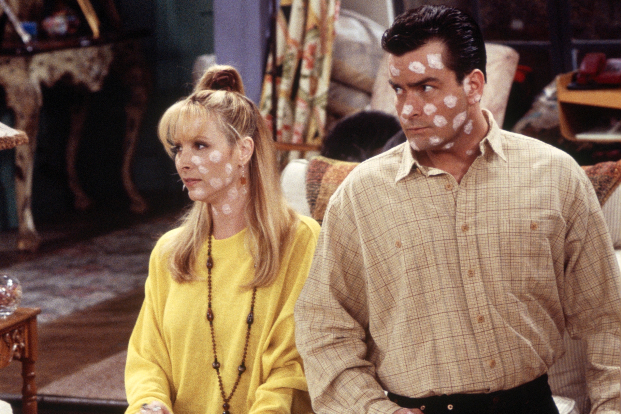 'Friends' Lisa Kudrow as Phoebe Buffay and Charlie Sheen as Ryan. Kudrow is wearing a yellow outfit and Sheen is wearing a tan collared shirt. They both have white spots all over to cover their chicken pox.