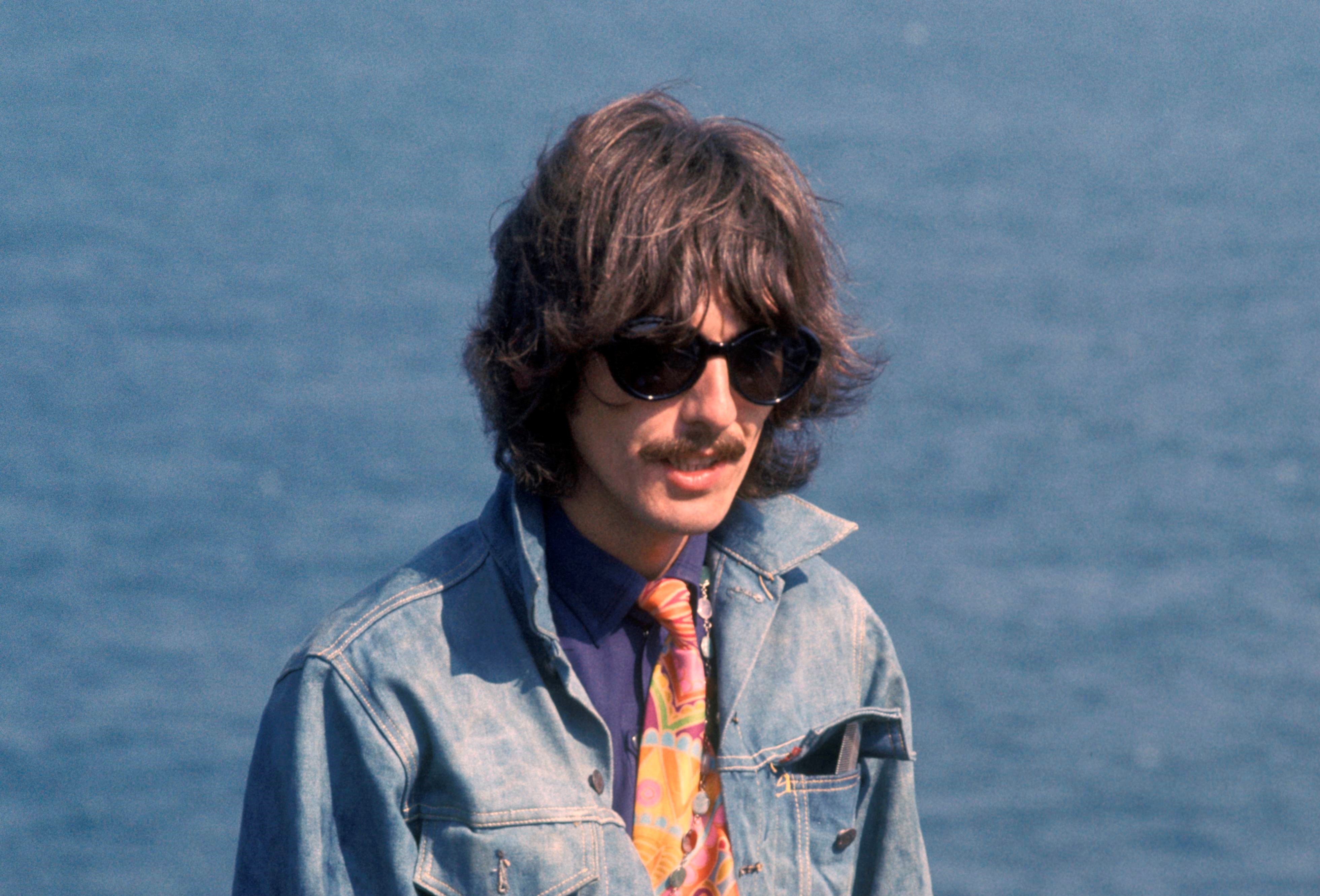 George Harrison wears a denim jacket and stands in front of water. 