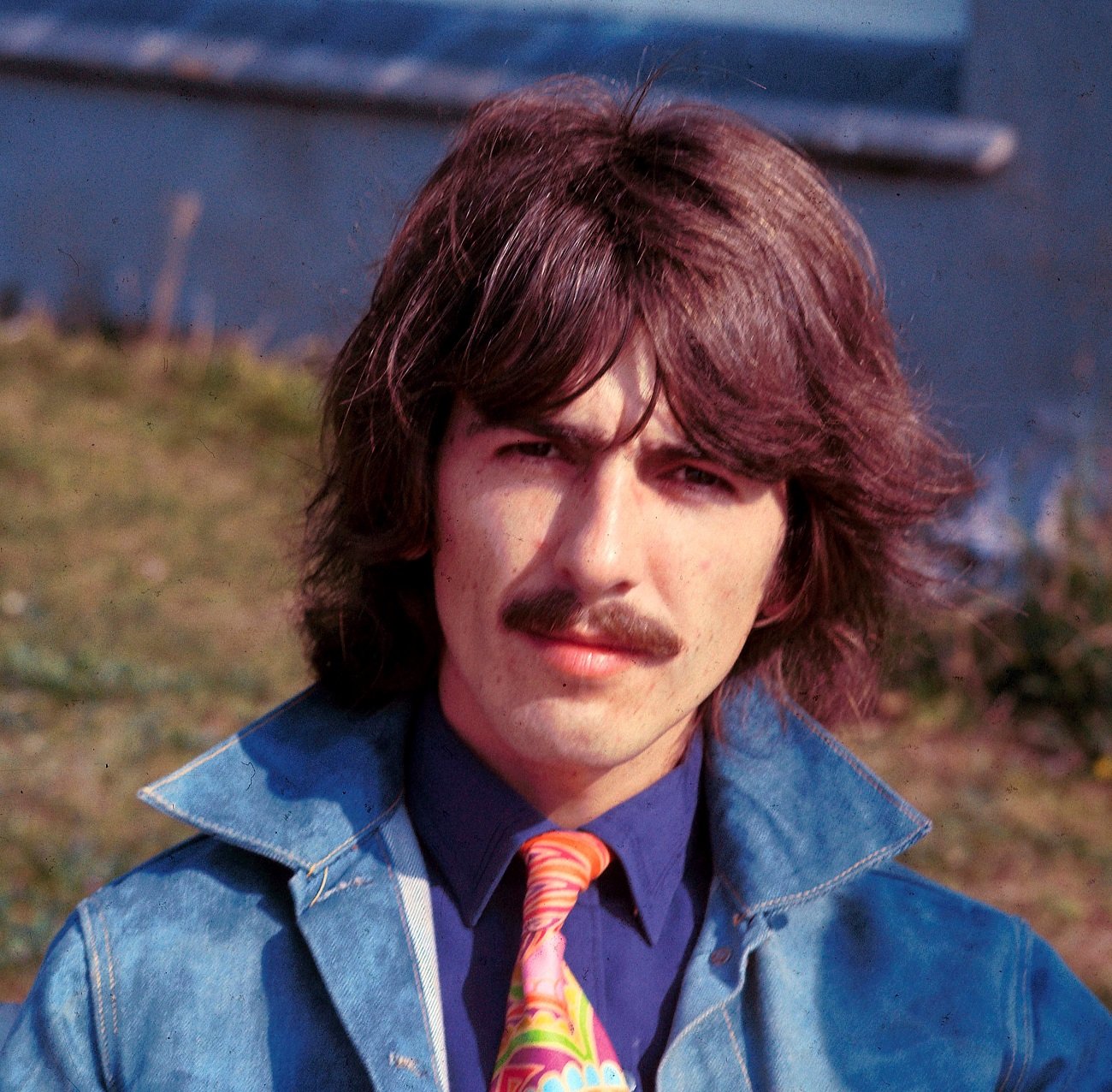 George Harrison wears a denim jacket and tie and poses near water.