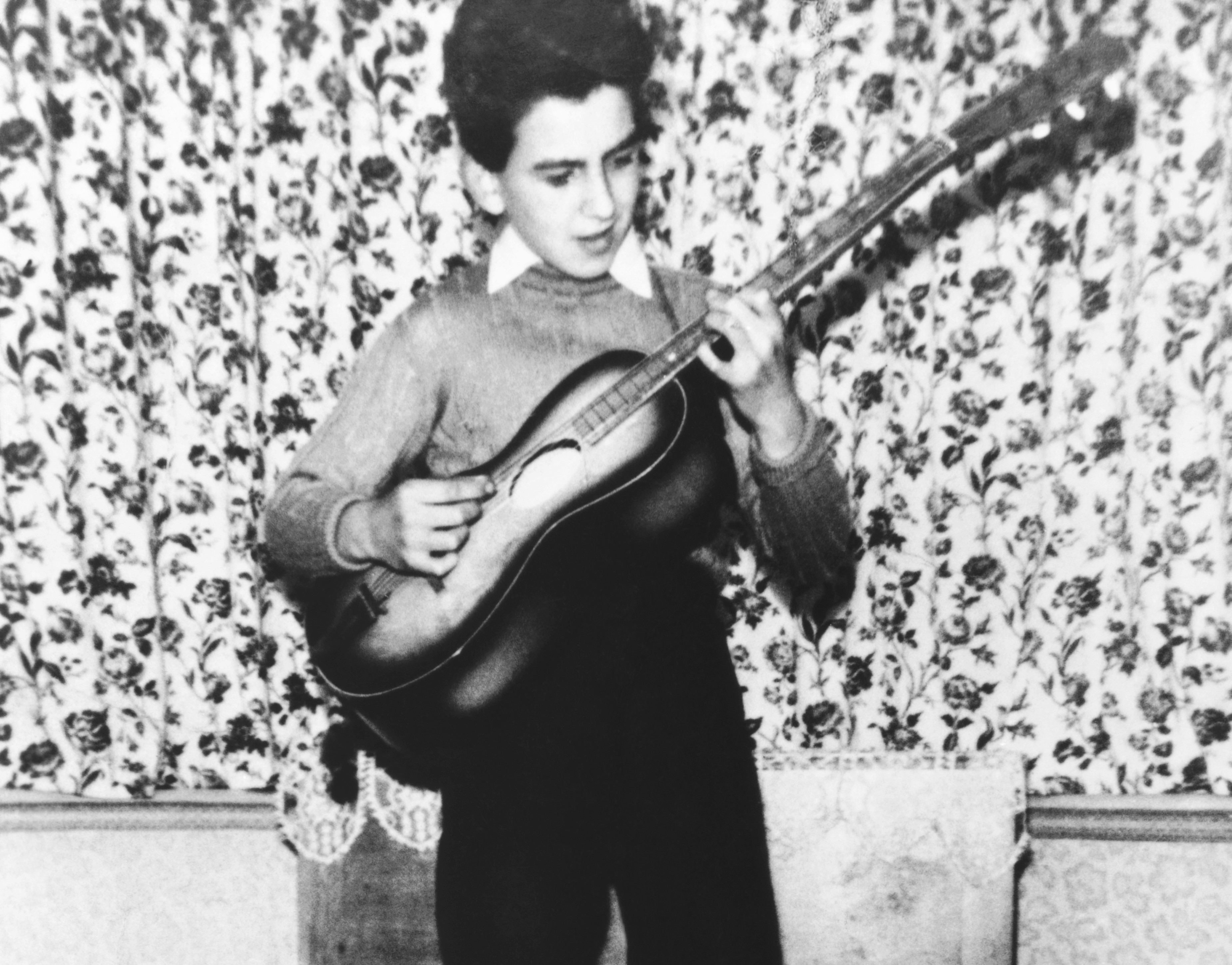 A black and white picture of a young George Harrison playing guitar in his family home. 