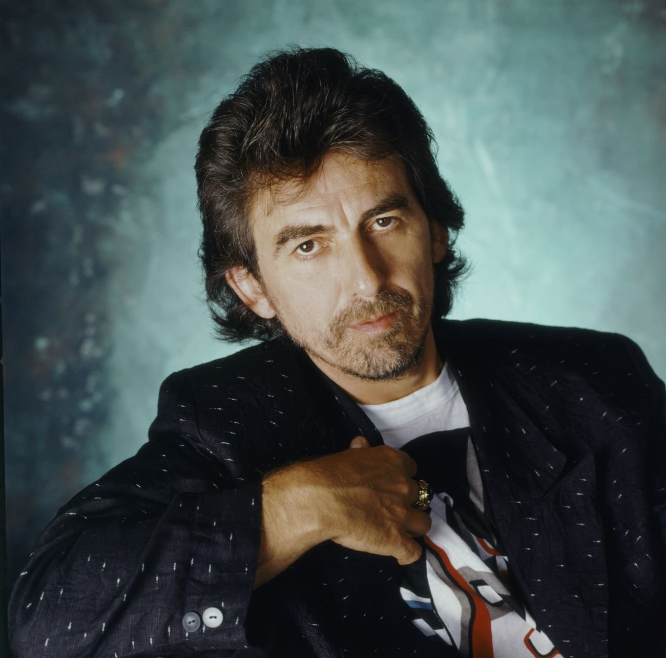 George Harrison leans over the side of his chair against a blue background.