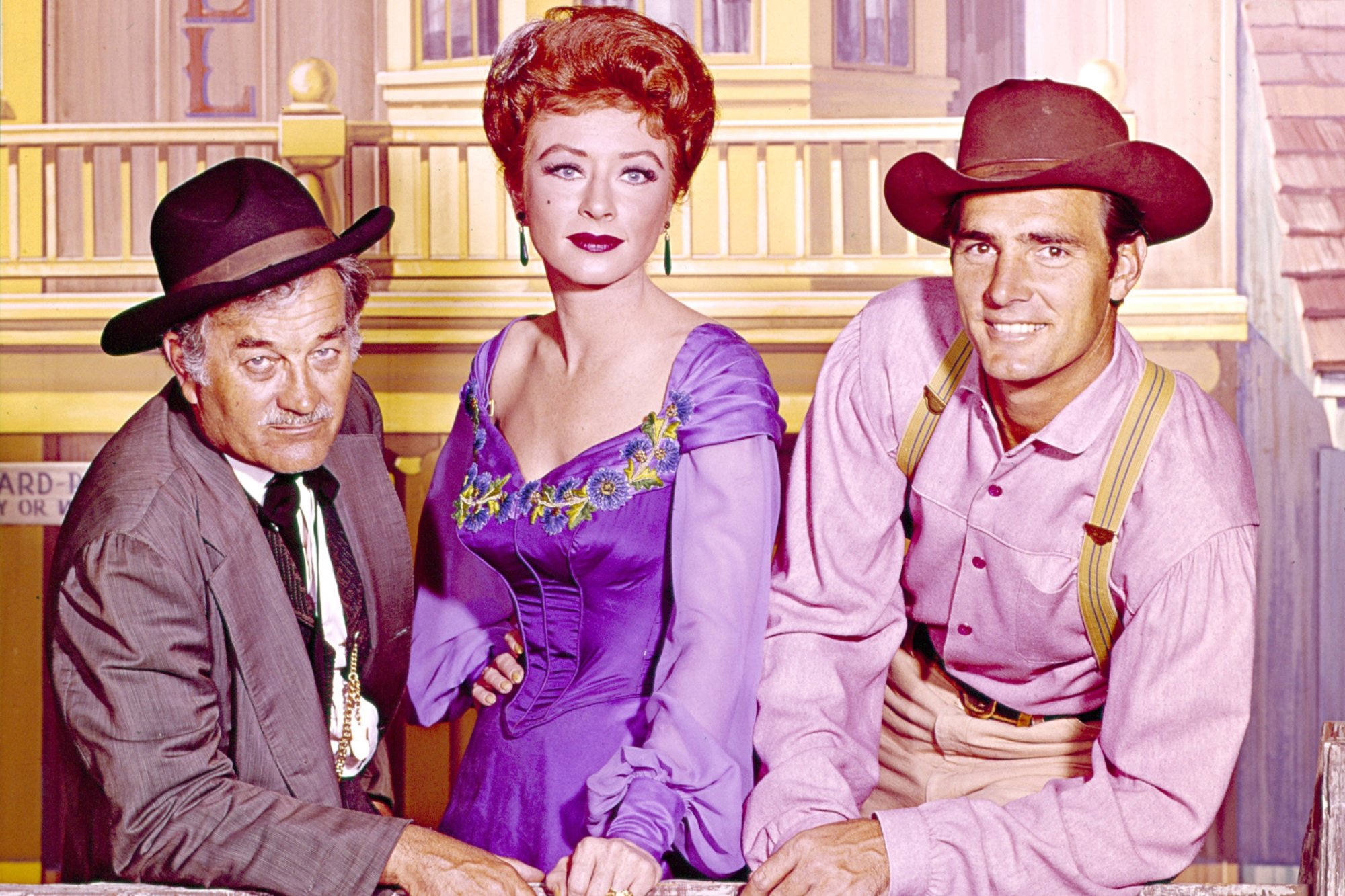 'Gunsmoke' Milburn Stone as Dr. Galen Adams, Amanda Blake as Kitty Russell, and Dennis Weaver as Chester Goode wearing Western clothes while leaning on a wooden fence. The hotel is the backdrop.