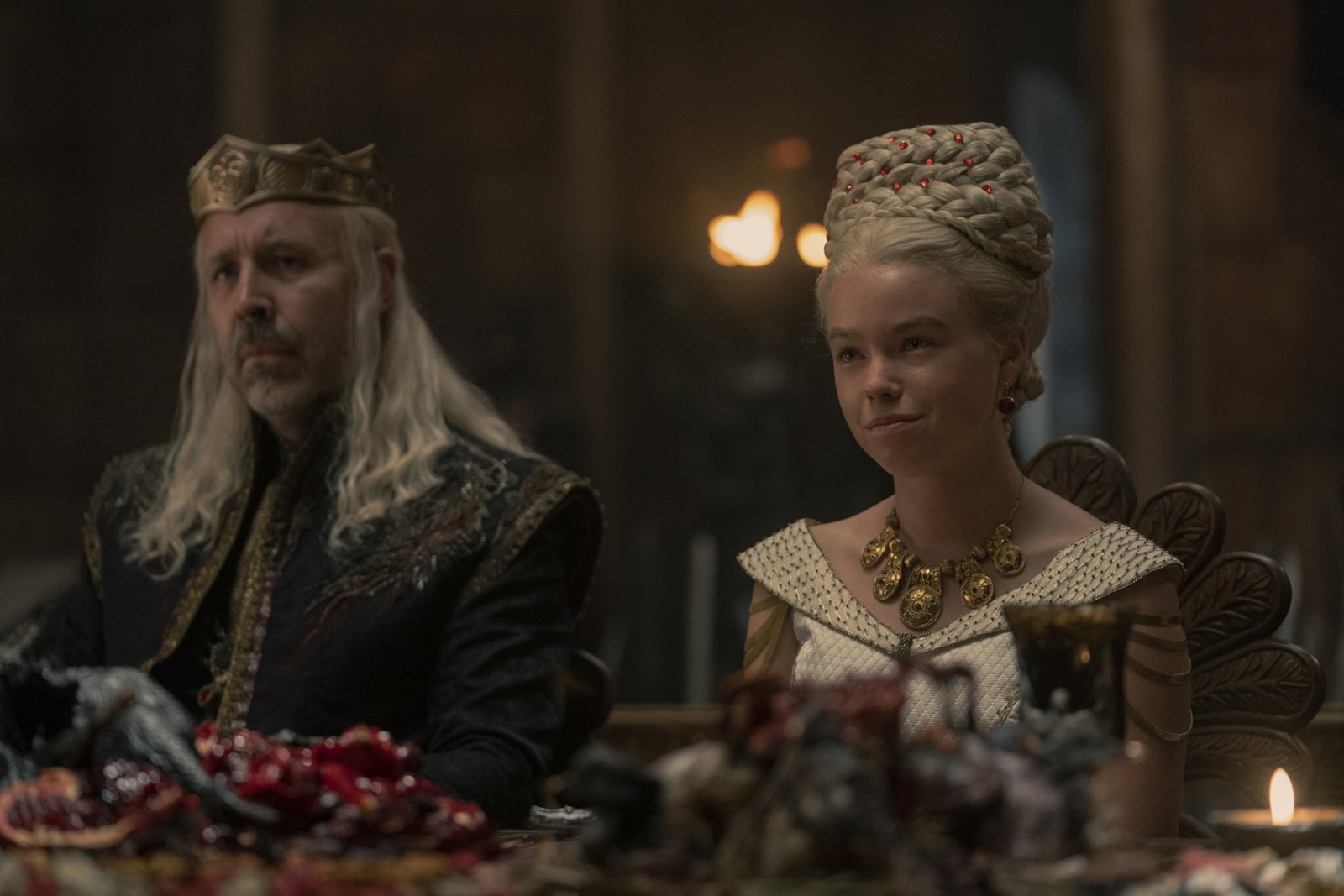 Paddy Considine and Milly Alcock as King Viserys and Princess Rhaenyra in 'House of the Dragon' Episode 5. They're both wearing finery, and Viserys has his crown on. They're sitting next to one another at a table.