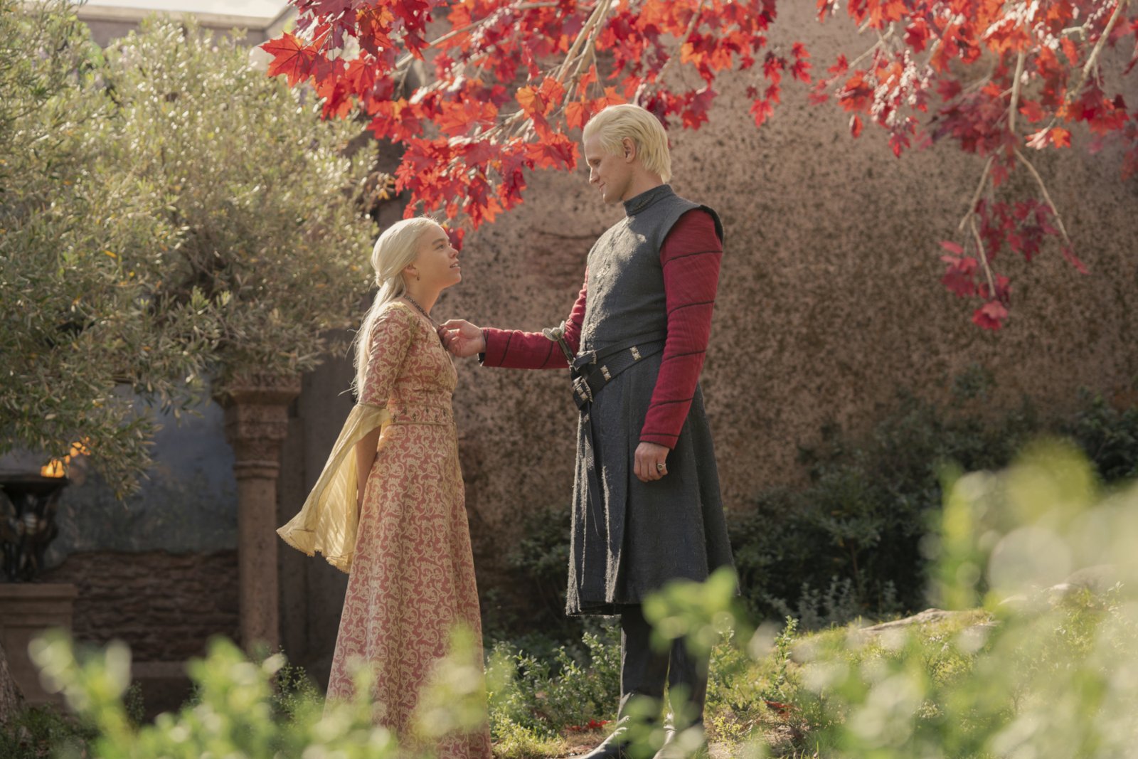 Milly Alcock and Matt Smith as Rhaenyra and Daemon Targaryen in 'House of the Dragon' for our article about episode 5. They're standing beneath a tree with red petals, and Daemon is holding the necklace around Rhaenyra's neck.