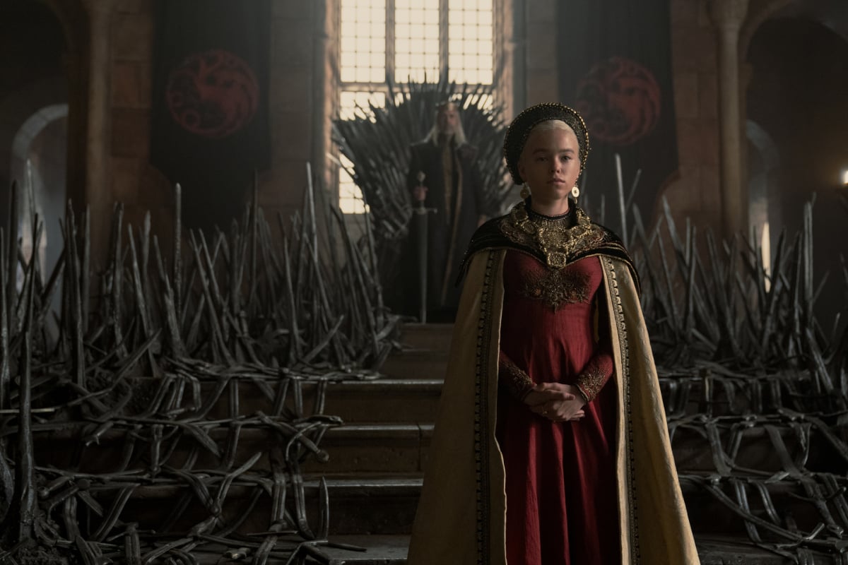 House of the Dragon costume designer Jany Temime created this look for Rhaenyra's coronation day. She wears a red dress, gold cape, and headpiece and stands in front of her father who is by the Iron Throne.