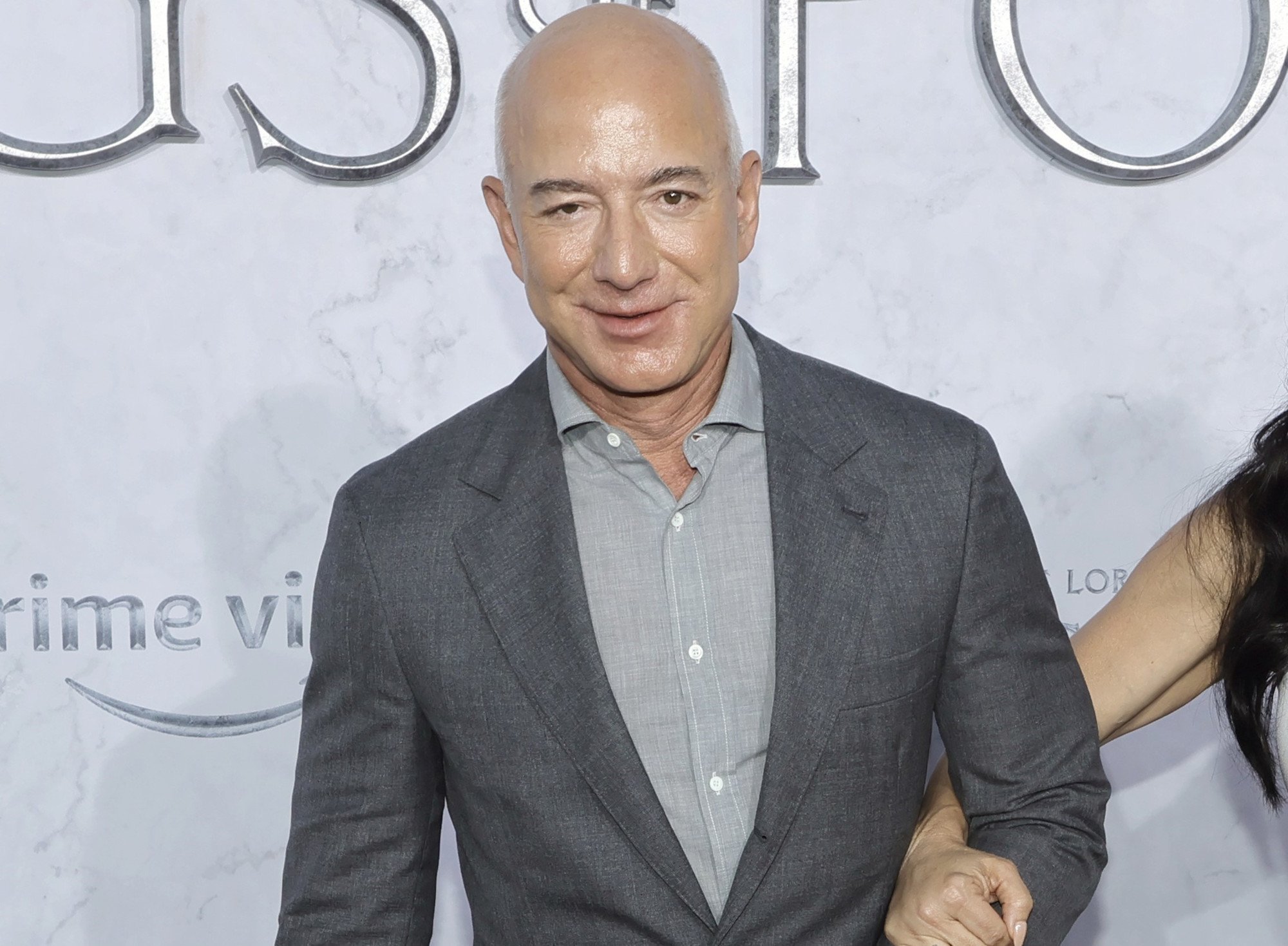 Jeff Bezos at the premiere for 'The Lord of the Rings: The Rings of Power.' He's wearing a grey shirt, darker grey jacket, and holding someone's arm.