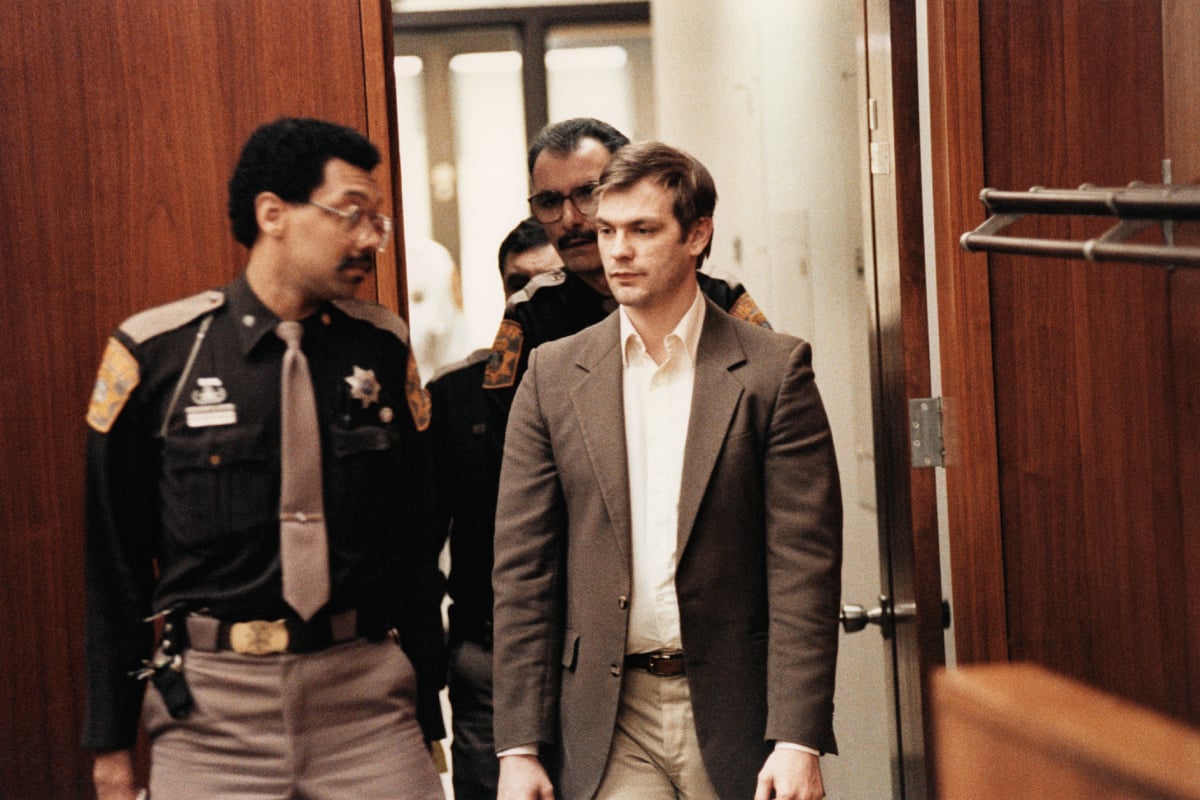 Jeffrey Dahmer is escorted into the courtroom by police. He wears a white button-down shirt and black suit jacket.