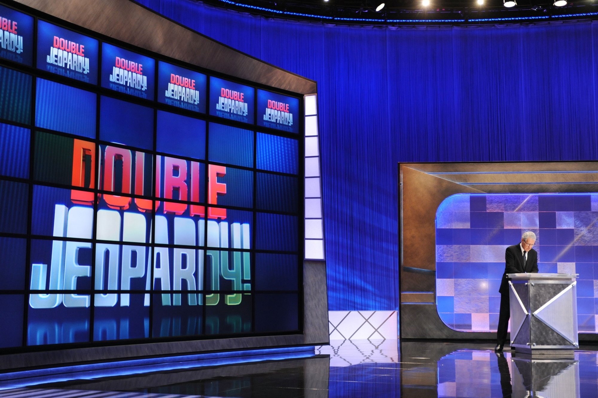 「危険！」 コマーシャルの前にショーをリードするアレックス・トレベック。 彼は、Double Jeopardy ボードを後ろにして、スーツを着て表彰台に立っています。