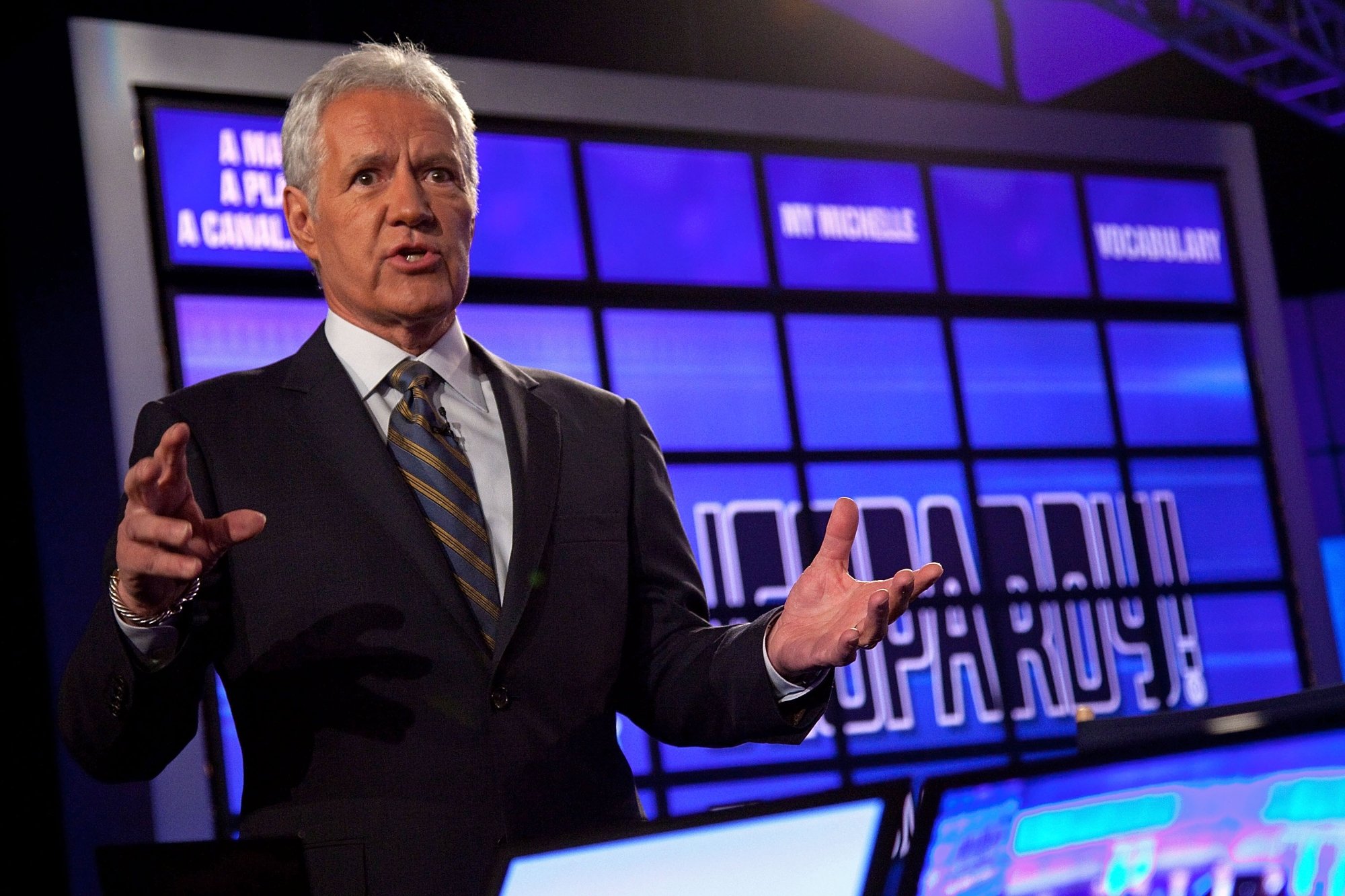 'Jeopardy!' Alex Trebek, speaking in front of the contestant podiums. He's wearing a suit in front of the digital board. He's talking with his hands in the air.