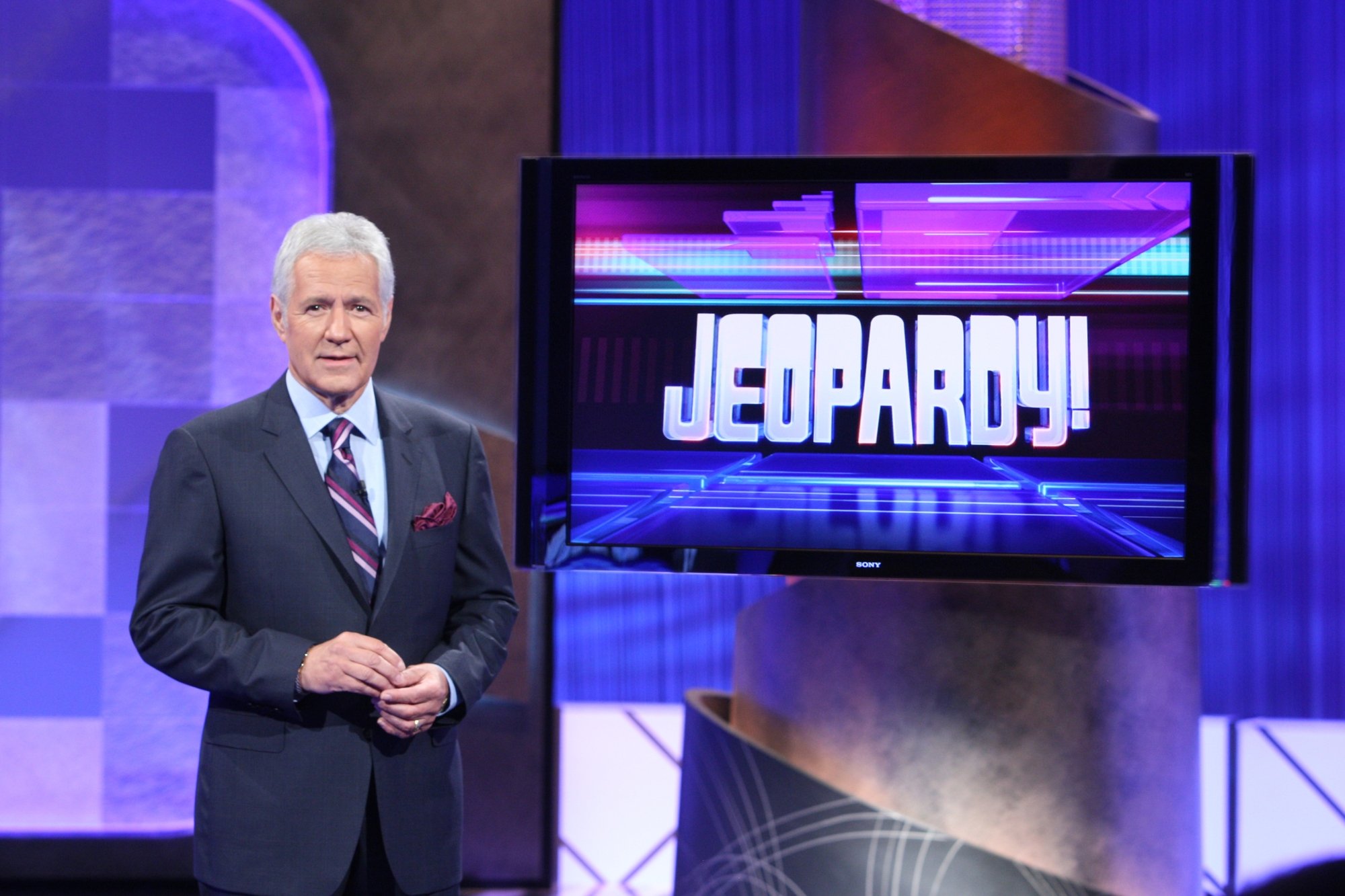 'Jeopardy!' Alex Trebek, who brought new buzzer rules. Trebek is wearing a suit, standing in front of a television with the 'Jeopardy!' logo on it