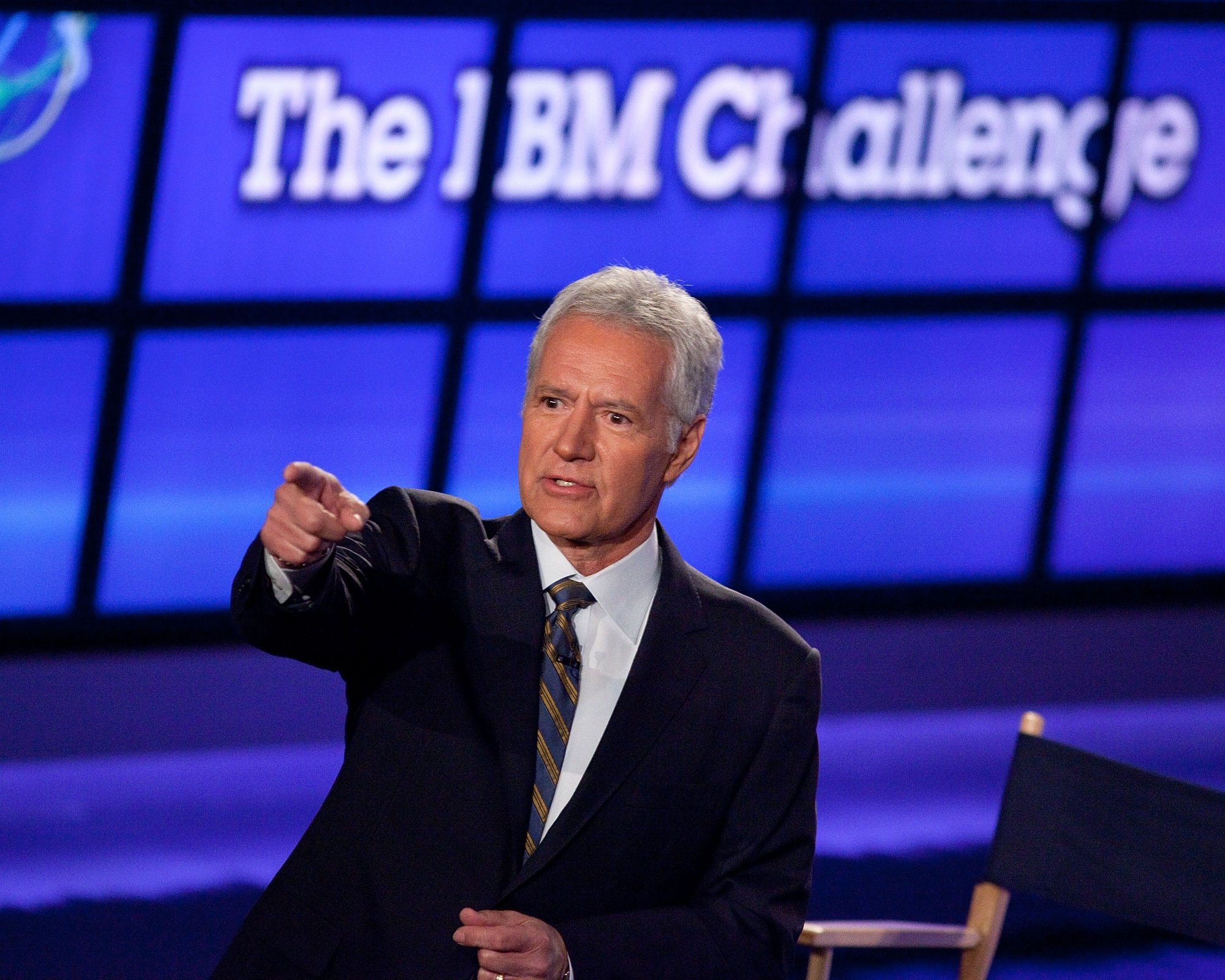 'Jeopardy! Alex Trebek, who enforced the most sacred rule. He's holding out his finger, pointing, while wearing a suit in front of blue screens.