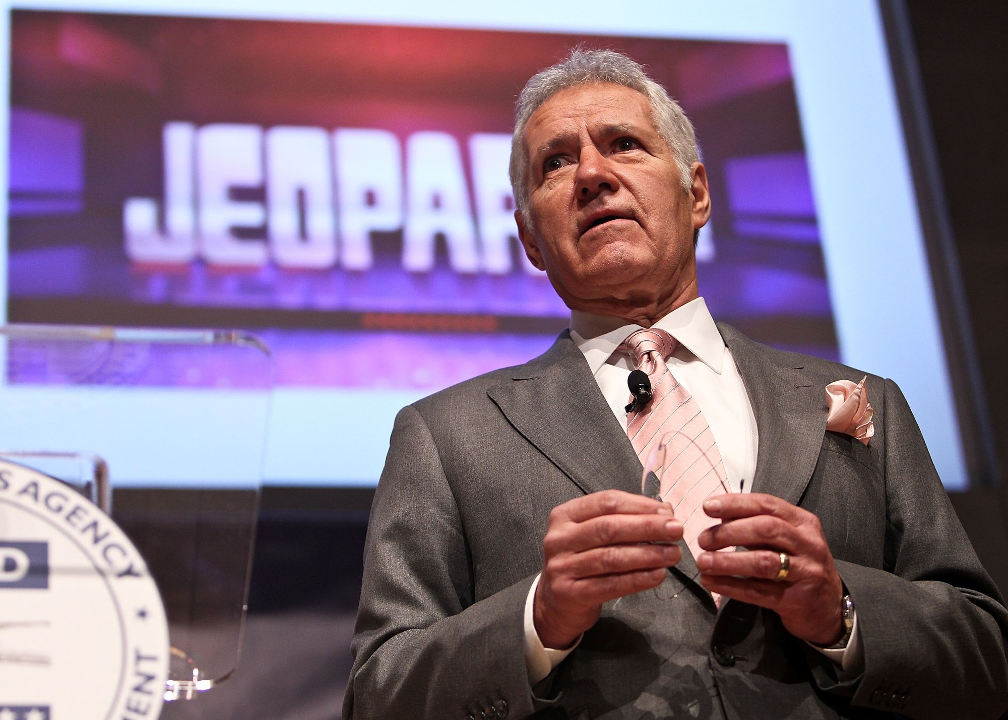 'Jeopardy!' Alex Trebek, who interacted with the contestant as a host. He's wearing a suit looking ahead with the show's logo on the screen behind him.