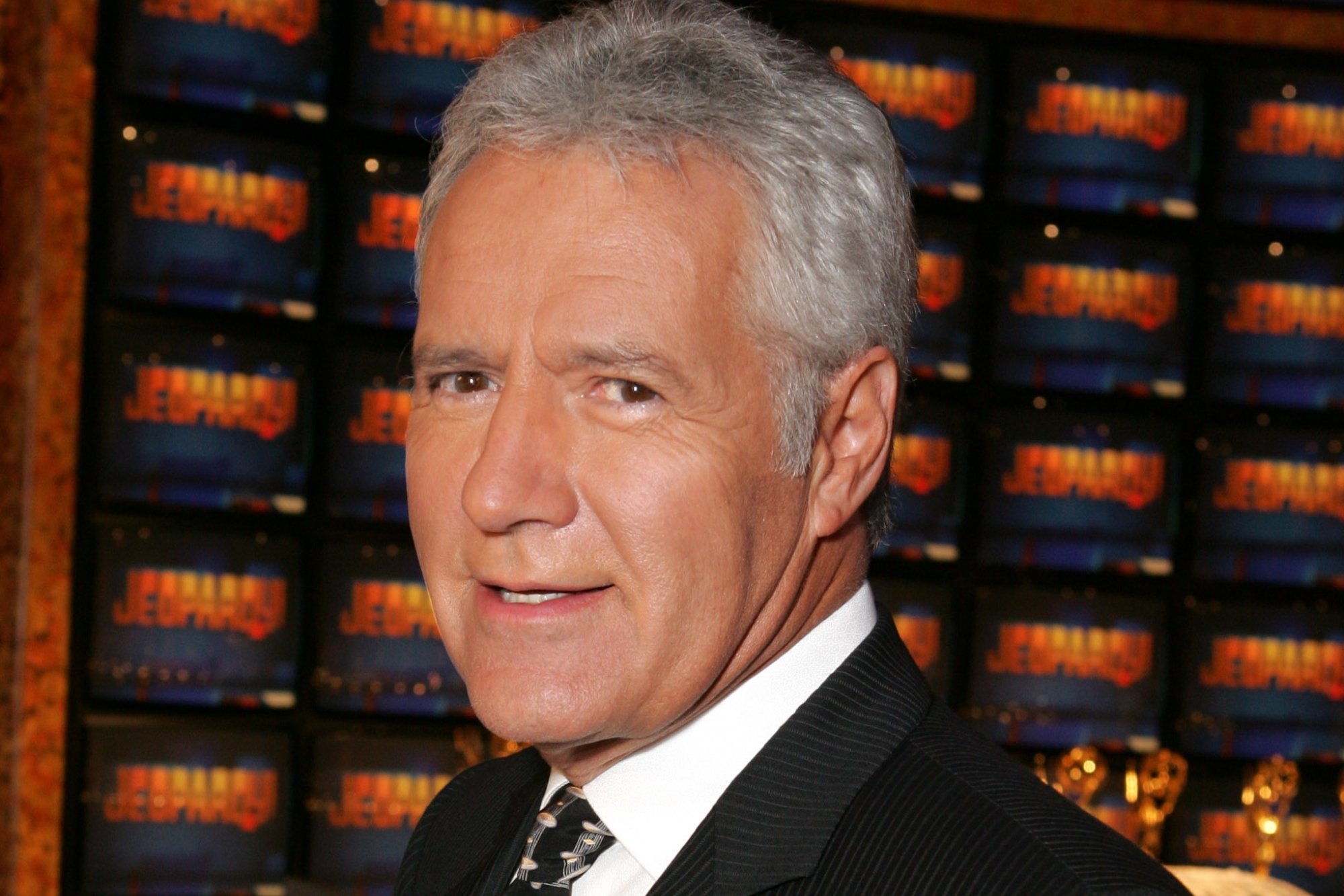 'Jeopardy!' host Alex Trebek who spoke with guests. He's wearing a dark suit jacket with a white collared-shirt underneath. He's standing in front of screens with the show's logo on them.