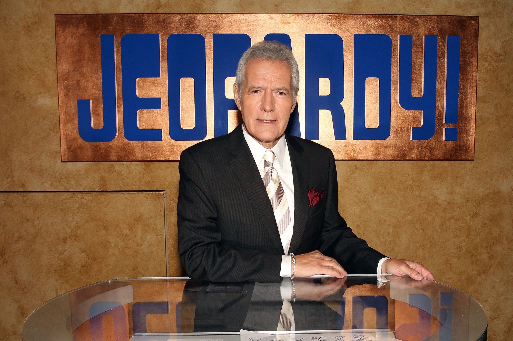 'Jeopardy!' host Alex Trebek posing against a table wearing a suit with the show's logo behind him