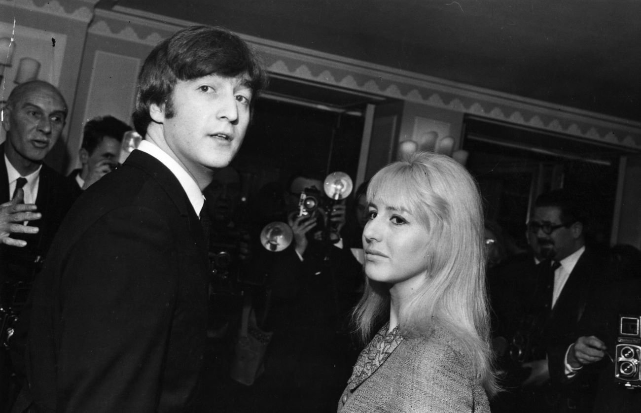 A black and white picture of John Lennon and Cynthia Lennon standing in a room full of photographers.