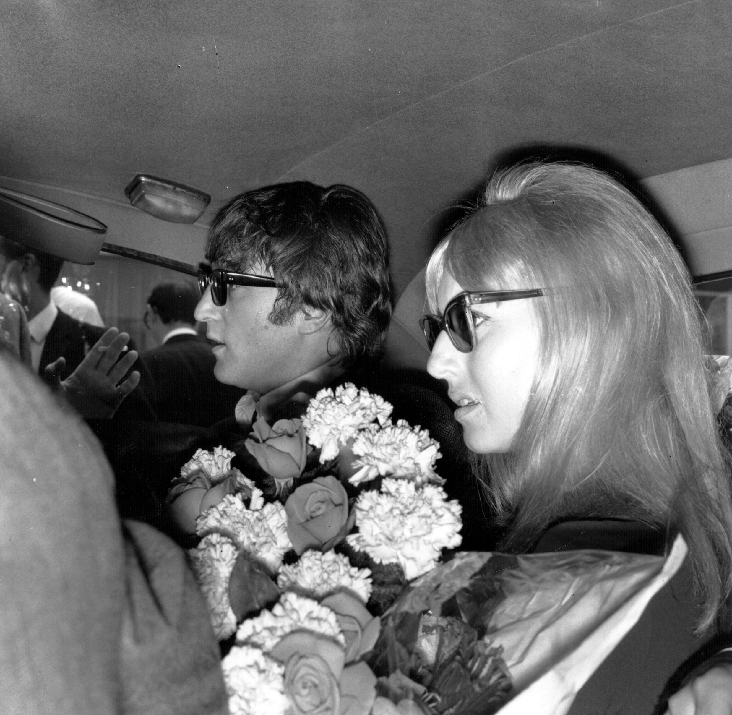 A black and white picture of John and Cynthia Lennon wearing sunglasses in the back seat of a car.