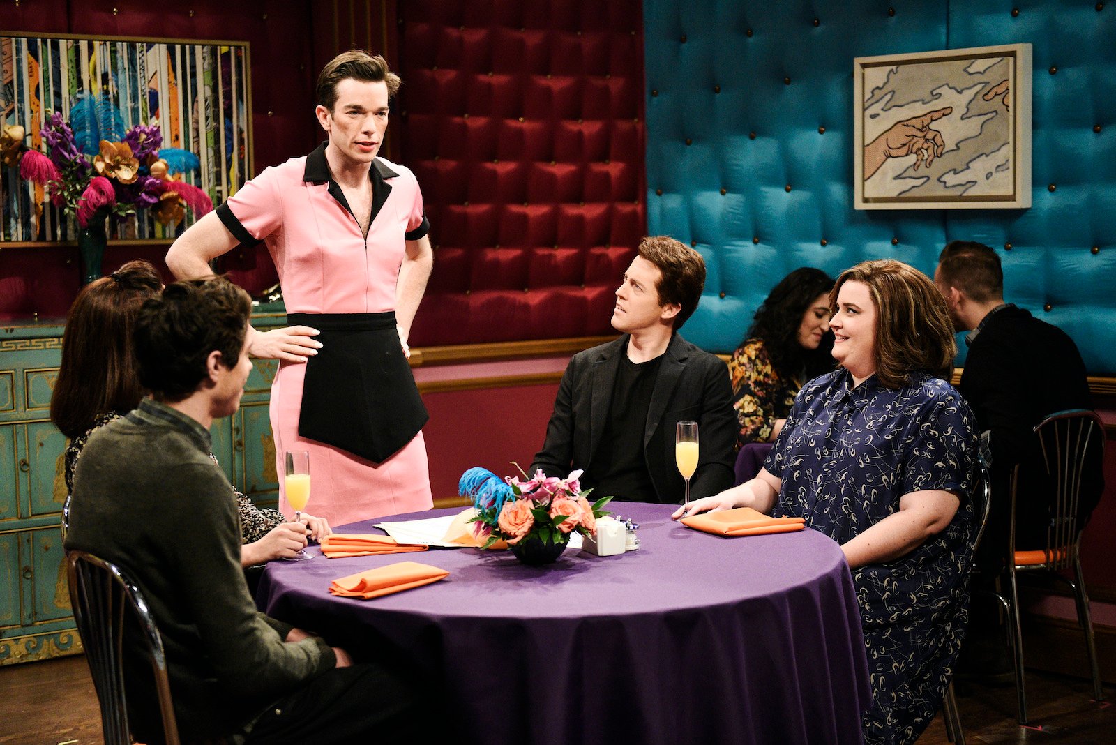 John Mulaney on 'SNL' dressed in a pink waitress costume stands next to a table of people with hands on hips. 