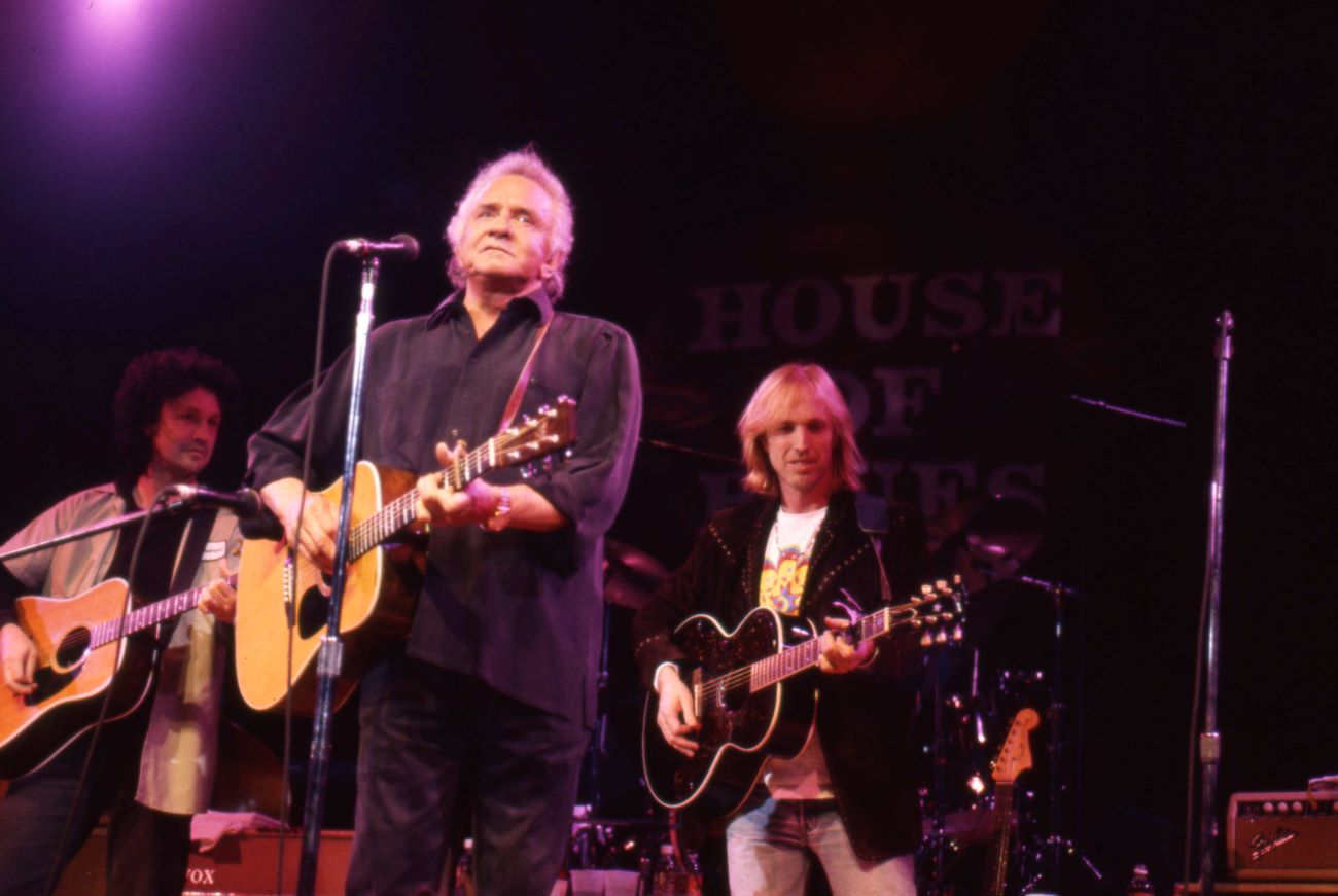 Johnny Cash holds a guitar and stands in front of a microphone. Tom Petty holds a guitar and stands behind him.