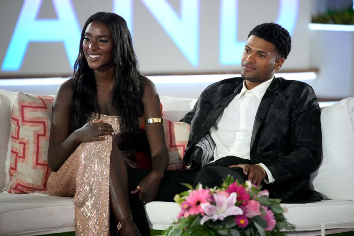 Zeta Morrison and Timmy Pandolfi, who appear during the 'Love Island USA' Season 4 reunion, sit on a white couch. Zeta wears a sparkly gold dress. Timmy wears a black suit over a white button-up shirt and black pants.