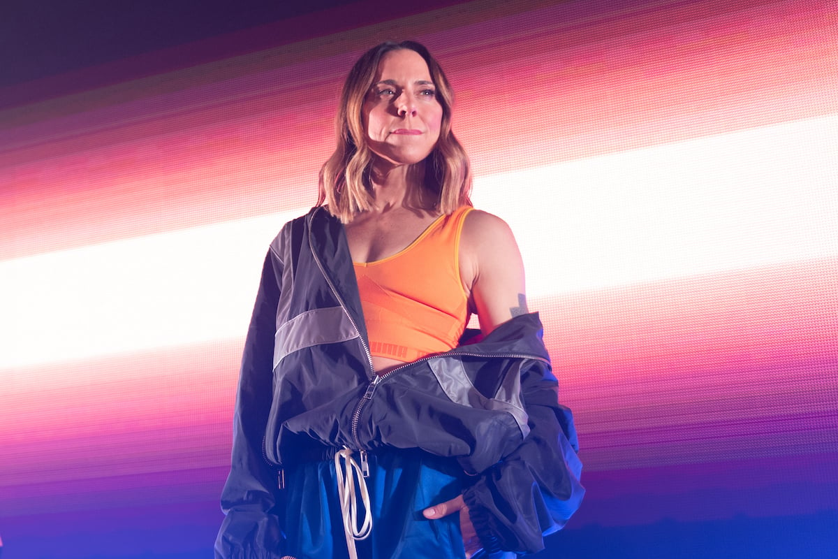 Mel C of the Spice Girls performing on stage wearing an orange top against a bright backdrop.