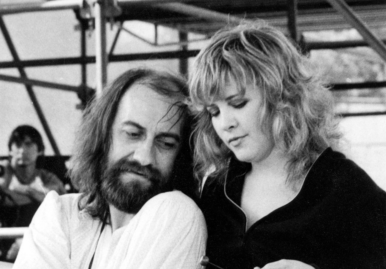 A black and white picture of Mick Fleetwood sitting and Stevie Nicks standing behind him.