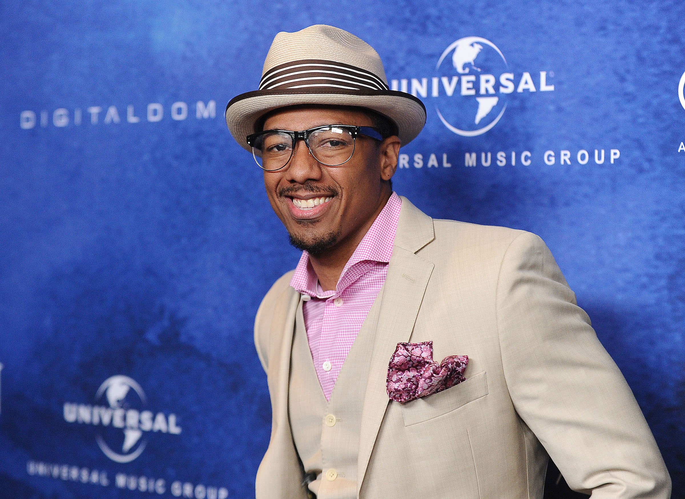 Nick Cannon, who has 9 children and counting, wearing a gray suit against a blue backdrop.