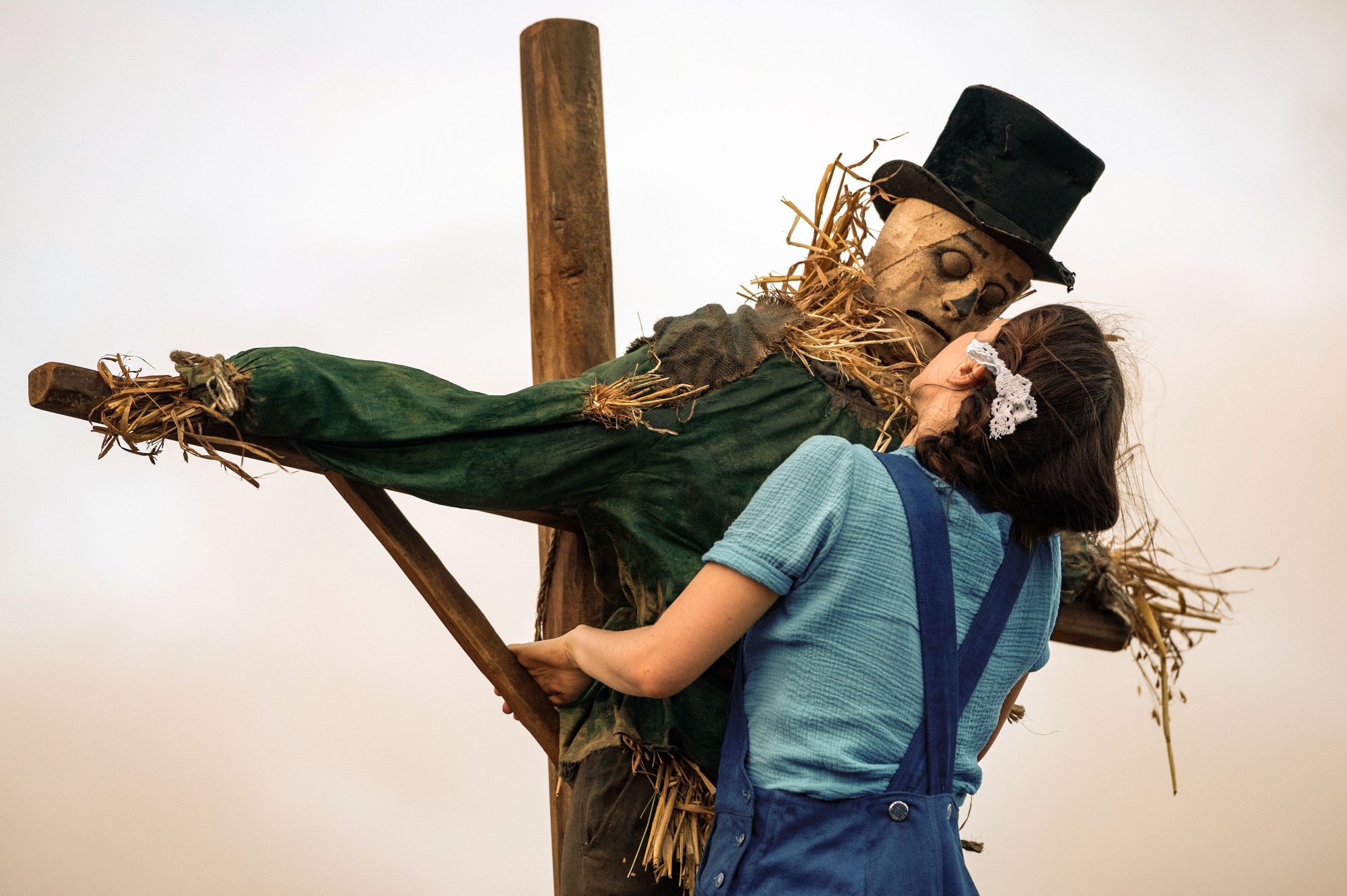 'Pearl' Mia Goth as Pearl with scarecrow climbing on top of a scarecrow cross. A top hat is on top of the scarecrow with her face close to the scarecrow's.