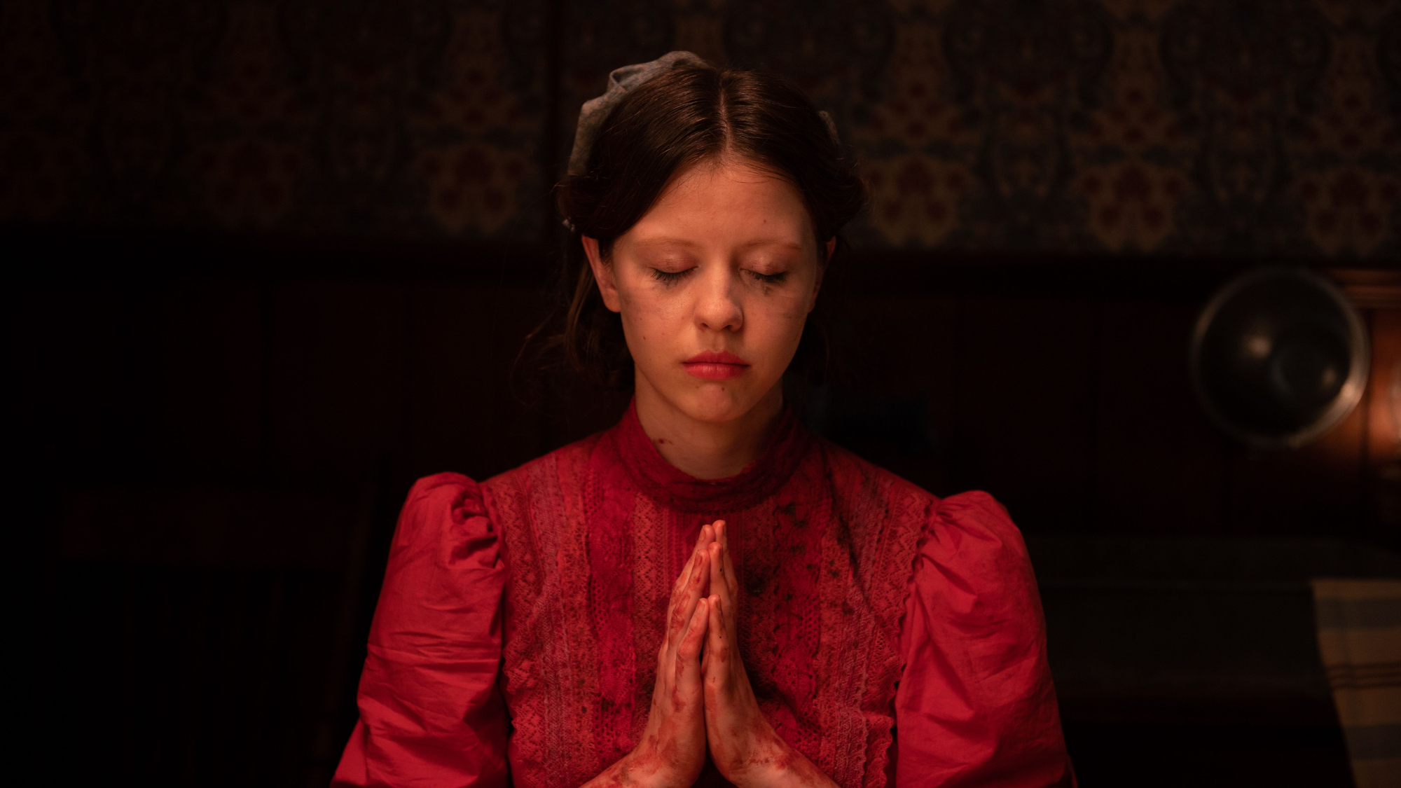 'Pearl' Mia Goth as Pearl holding her hands together to pray. She has her eyes closed, wearing a red dress, with her hands covered in blood.
