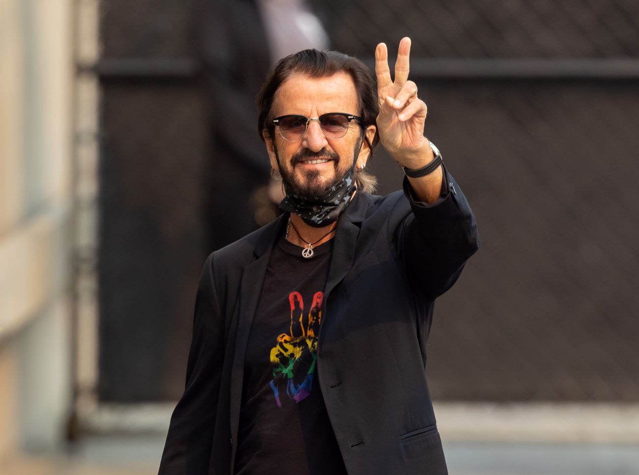 Ringo Starr wears a peace sign shirt and holds his hand in a peace sign.