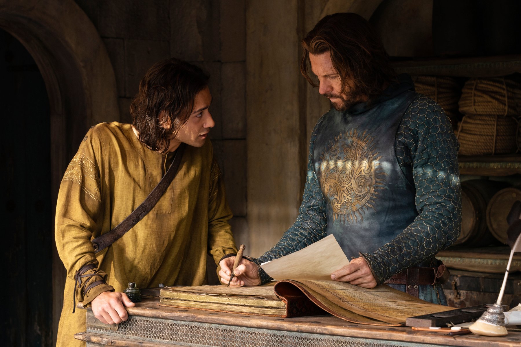 Maxim Baldry and Lloyd Owen as Isildur and Elendil in 'The Rings of Power' Episode 5 for our article about its Easter Eggs. Isildur is leaning over a table and talking to his father, and Elendil is writing on paper and listening.