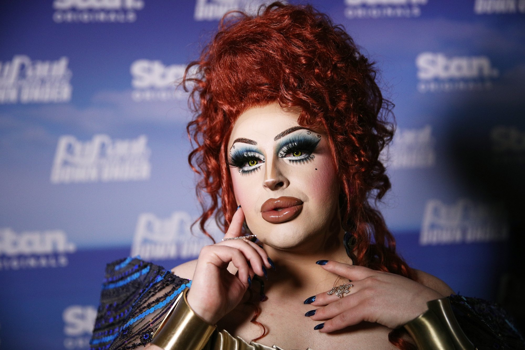 'RuPaul's Drag Race Down Under' Season 2 Hannah Conda posing with one hand near her neck and the other resting on her cheek. She's wearing red hair in front of a 'Drag Race' step and repeat.