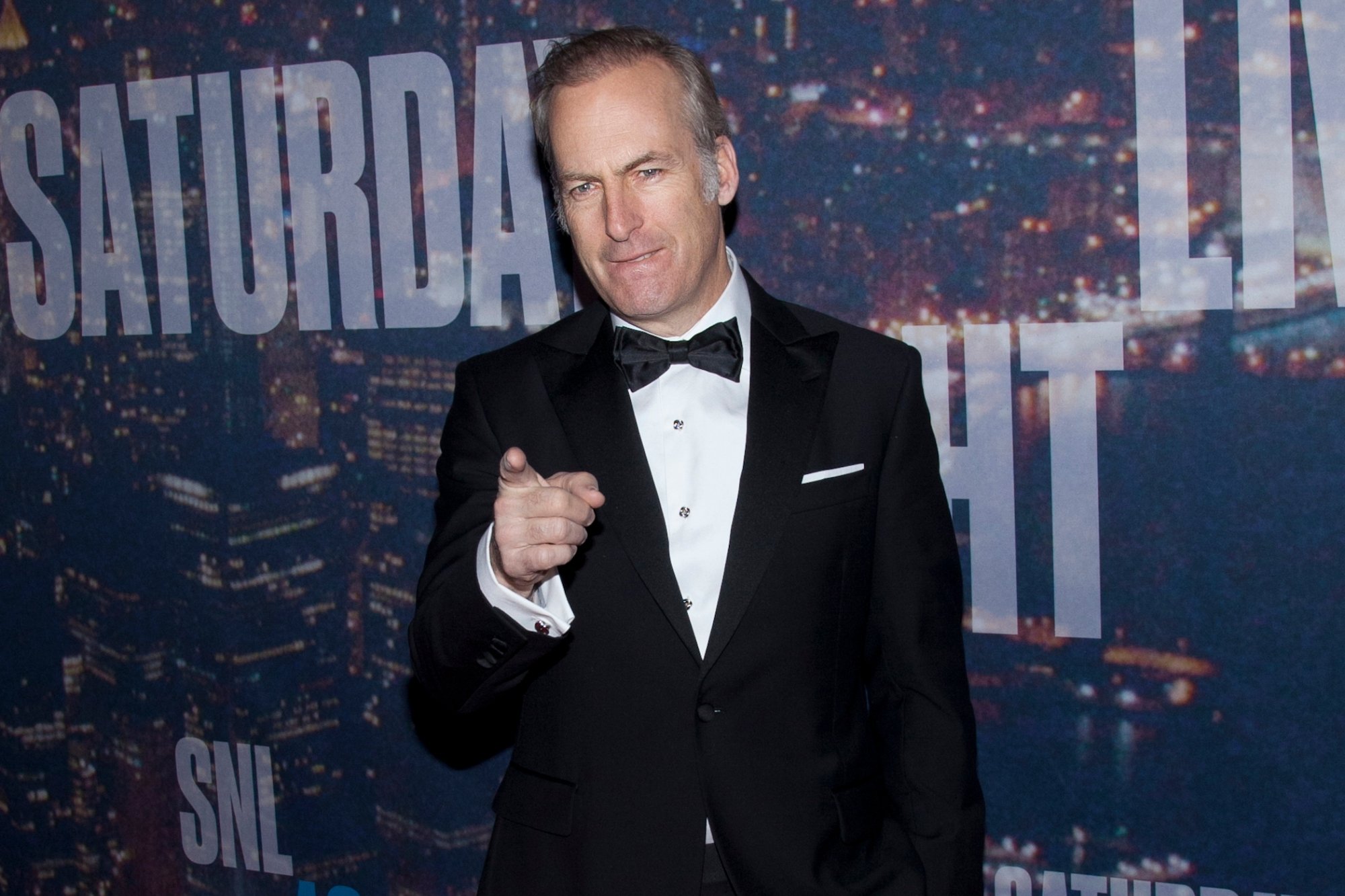 'Saturday Night Live' Bob Odenkirk wearing a tuxedo in front of 'SNL' step and repeat. He's pointing his finger out while his other hand is in his pocket.